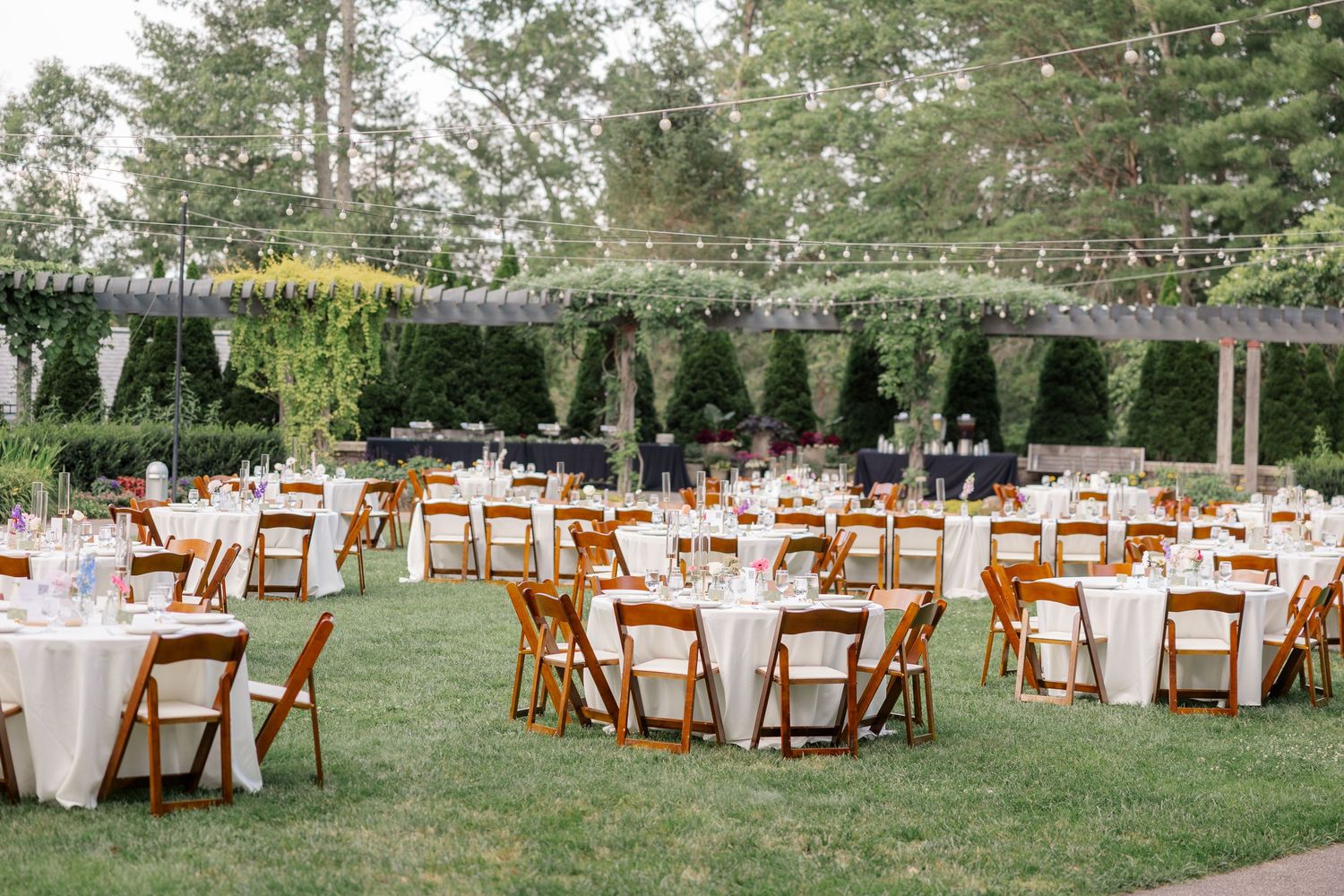 Outdoor wedding reception setup with wooden chairs and white tablecloths under tall trees.