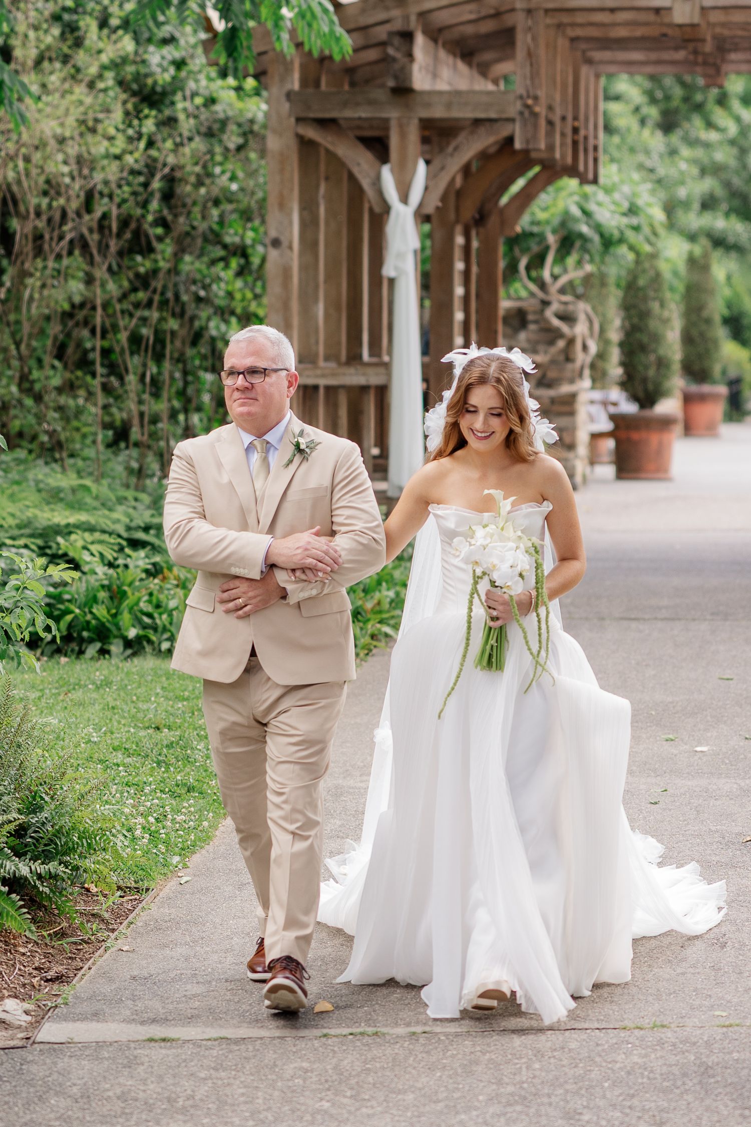 A bride in a flowing white gown walks with her father along a garden path.