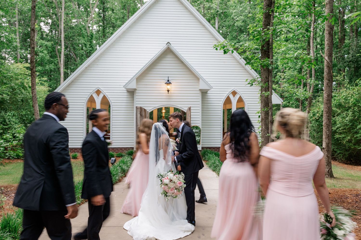 Wedding party in pink and black formal attire gathered outside a white chapel.