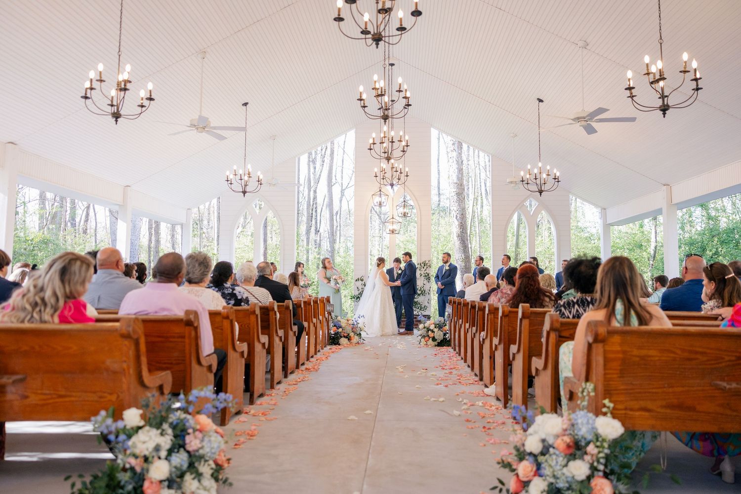 Church interior decorated with chandeliers and floral arrangements along wooden pews for a wedding ceremony.