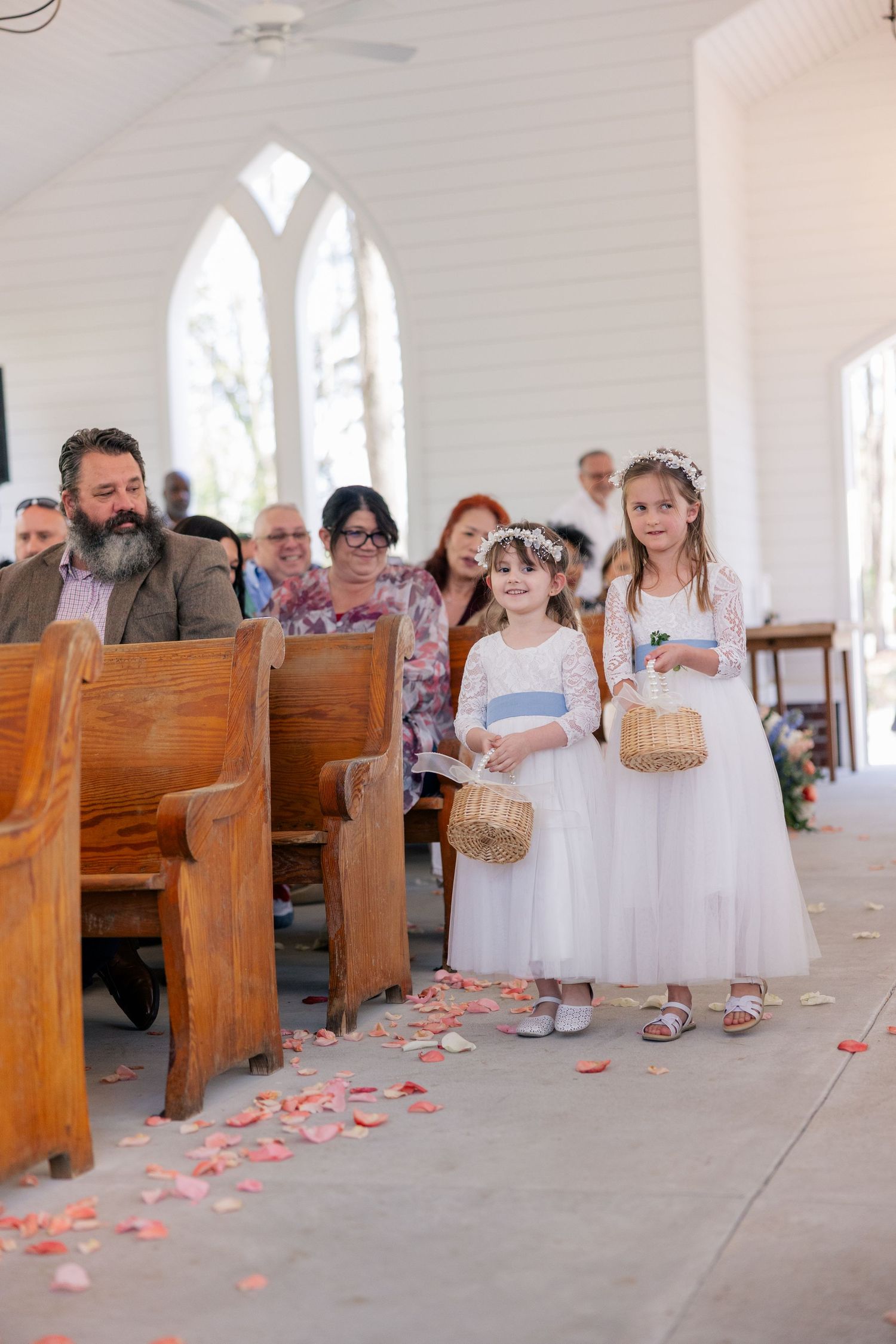 Young flower girls in white dresses walk down church aisle scattered with rose petals.