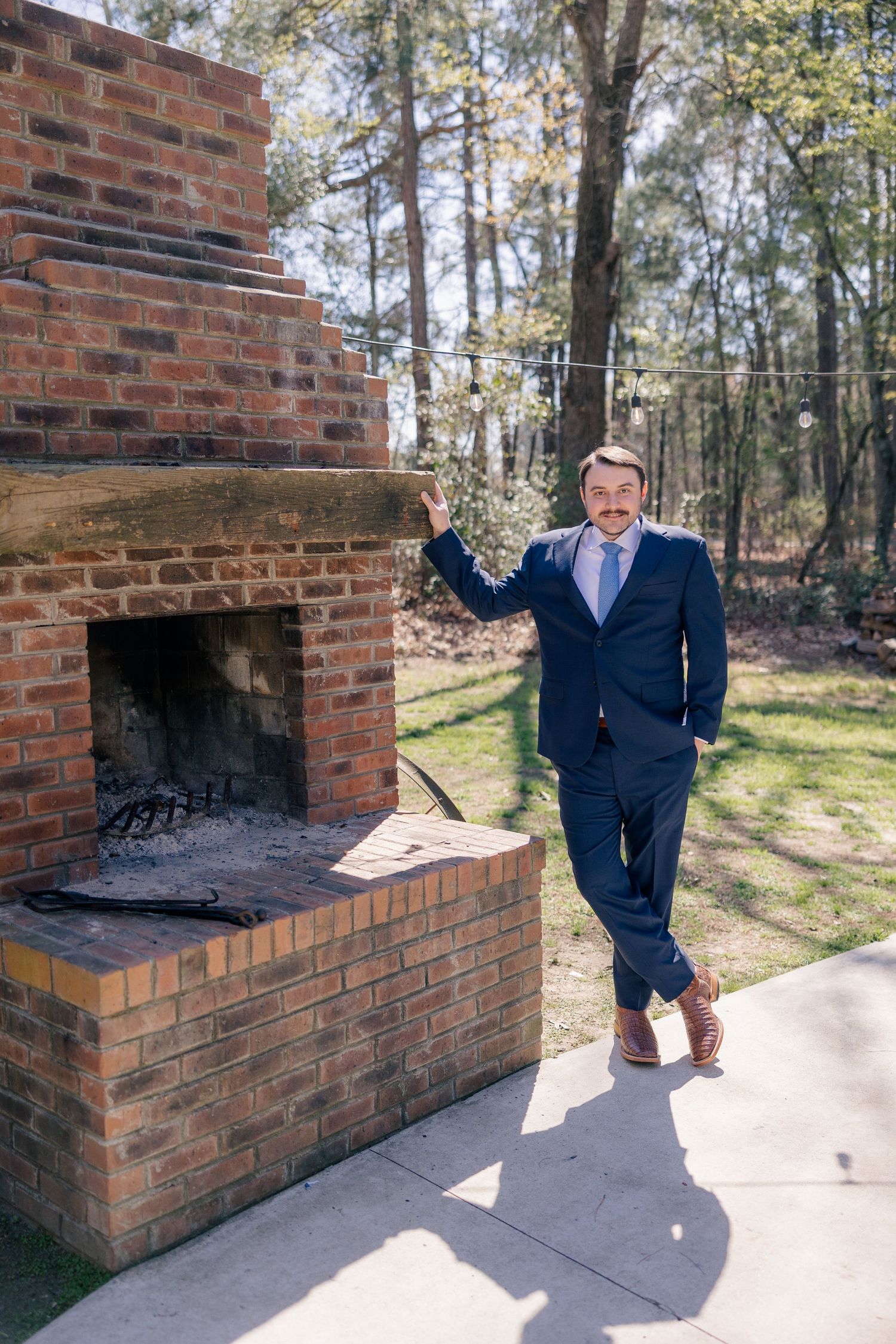 A person in a navy suit poses playfully near a brick outdoor fireplace.