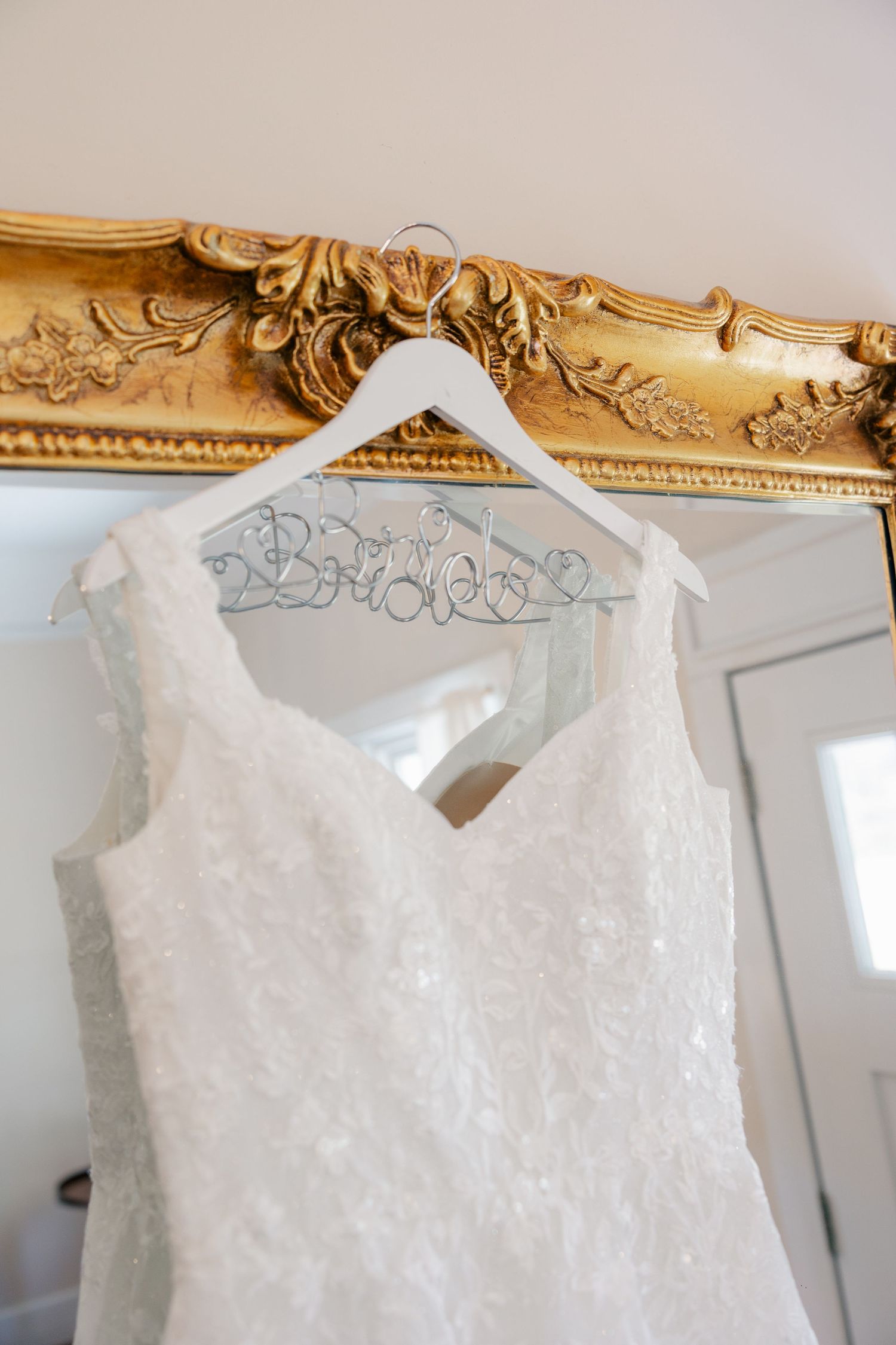 Elegant white lace wedding dress hangs from ornate gold frames in a bridal suite.