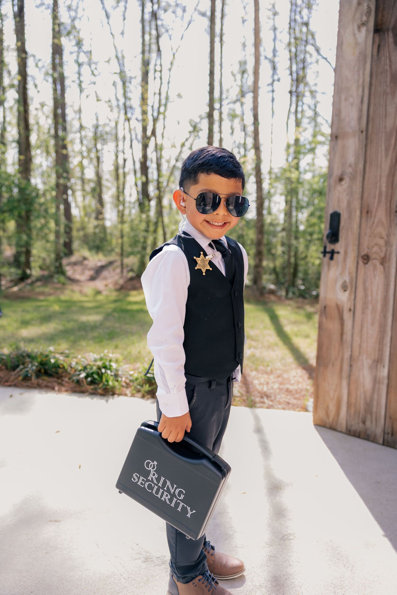 A young ring bearer in a black vest and sunglasses carries a wedding sign outdoors.