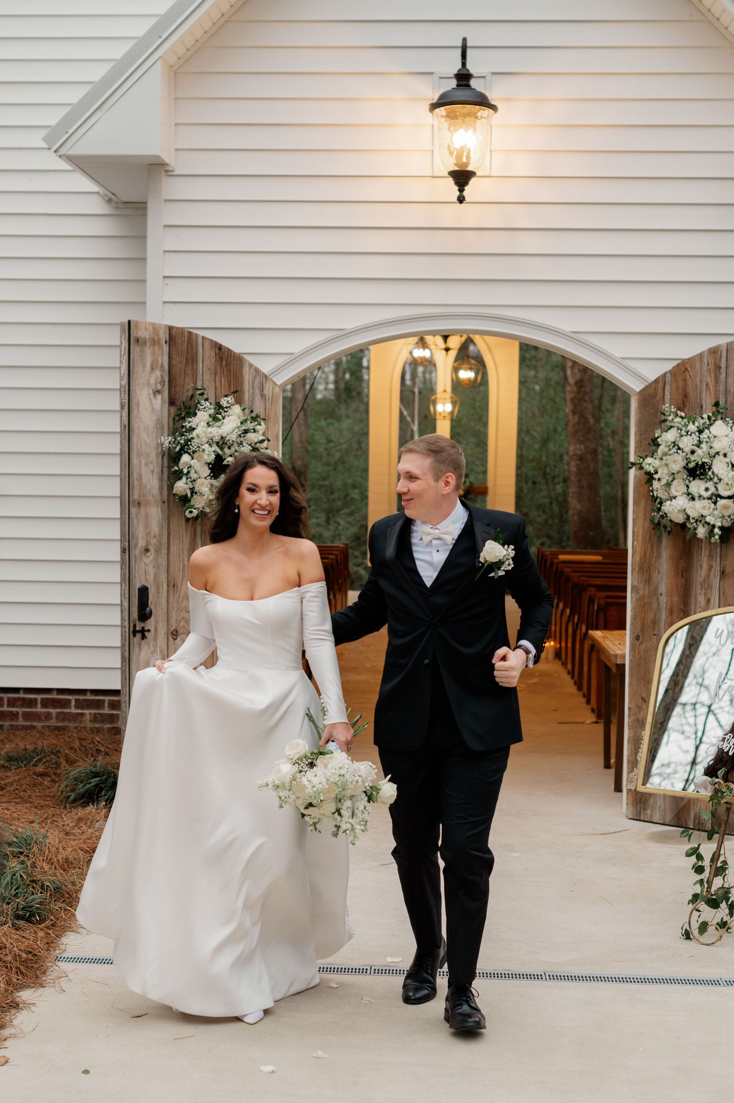 An elegant bride and escort walk through a white-painted covered walkway adorned with glowing lanterns and floral decor.