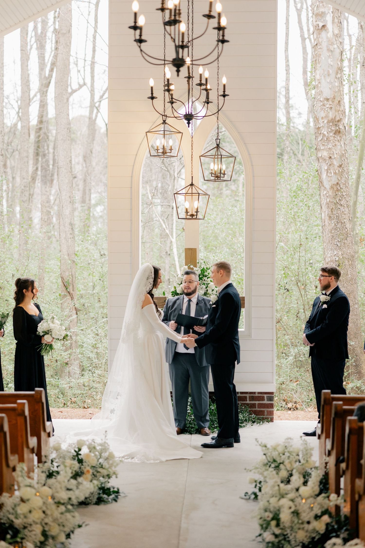 Light-filled chapel wedding ceremony with white floral decorations and elegant chandeliers.