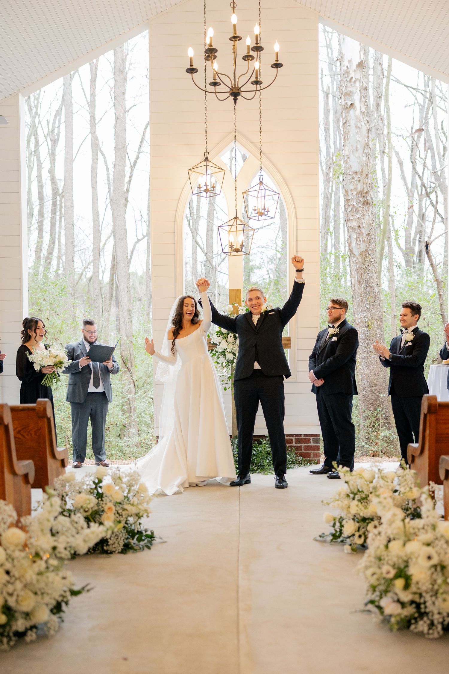 Newlyweds celebrate with raised arms at altar surrounded by white floral arrangements.