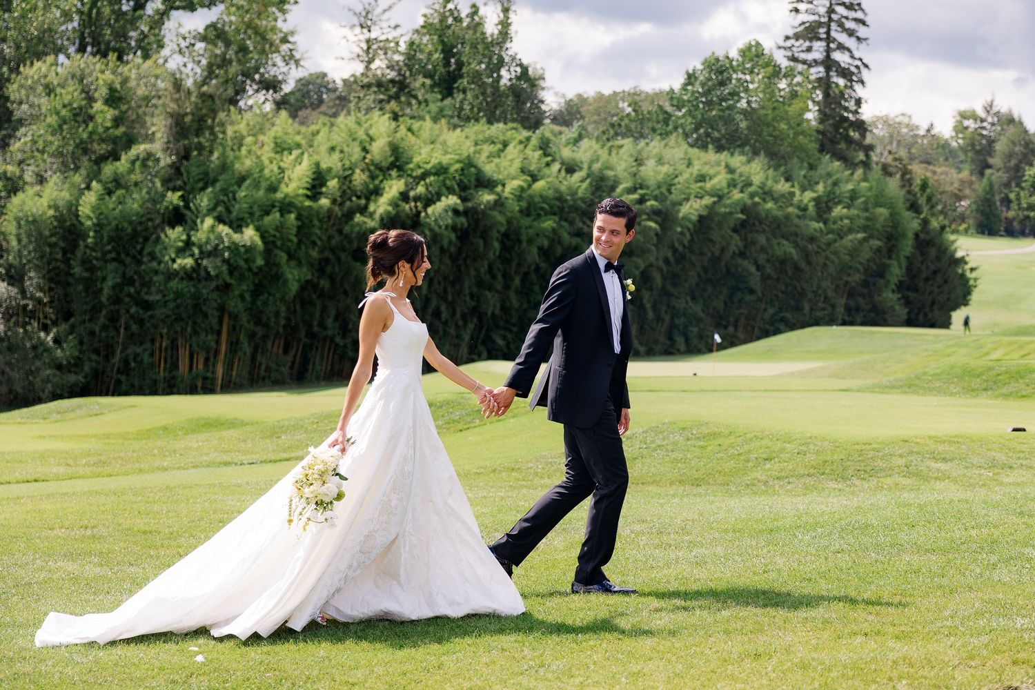 Bride and groom holding hands walking on lush green golf course lawn on their wedding day.