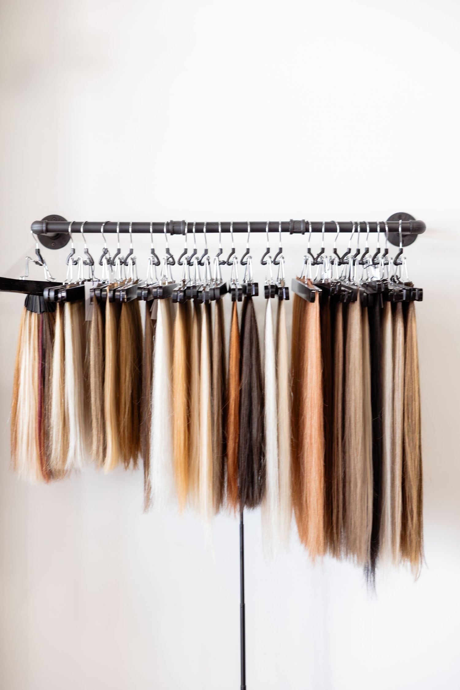 A display rack of hair extensions in various shades from blonde to dark brown hanging on silver clips against a white wall.