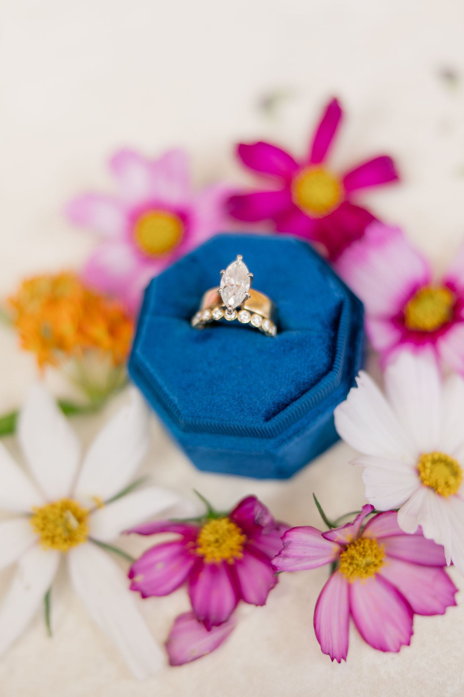 Marquise diamond engagement ring with wedding band in blue velvet ring box surrounded by colorful wildflowers.
