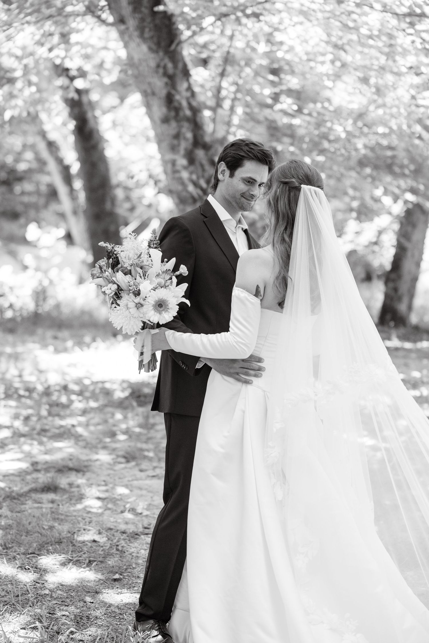 Black and white portrait of bride and groom embracing under trees, bride holding a bouquet with veil flowing.