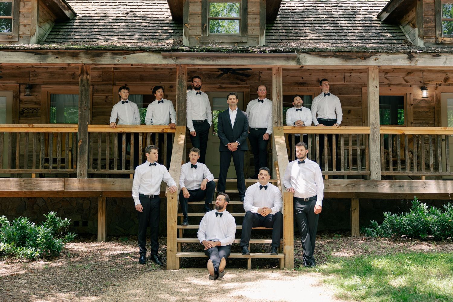 Groom and groomsmen in black tuxedos pose on rustic log cabin steps for a classic wedding party photo.