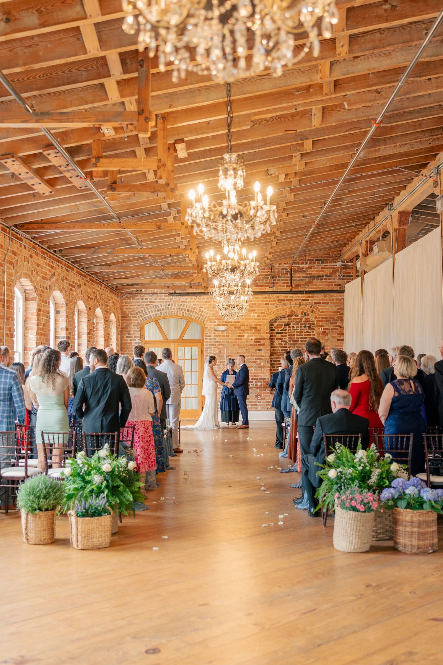 Guests stand in a rustic brick wedding venue with chandeliers and basket floral arrangements lining the aisle.
