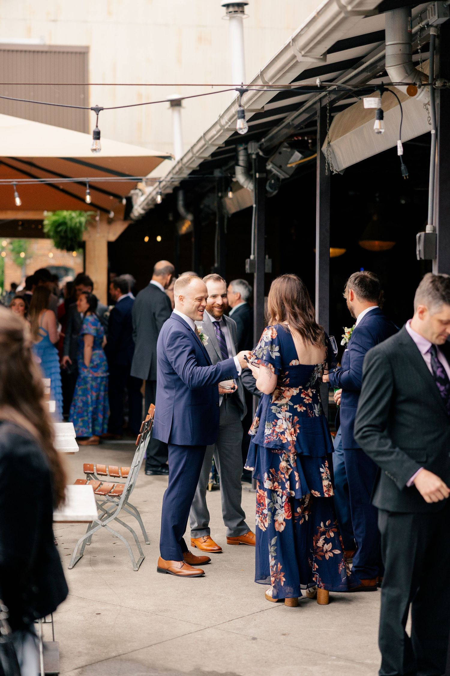 Wedding guests mingle outdoors on a patio under string lights during a cocktail hour reception.