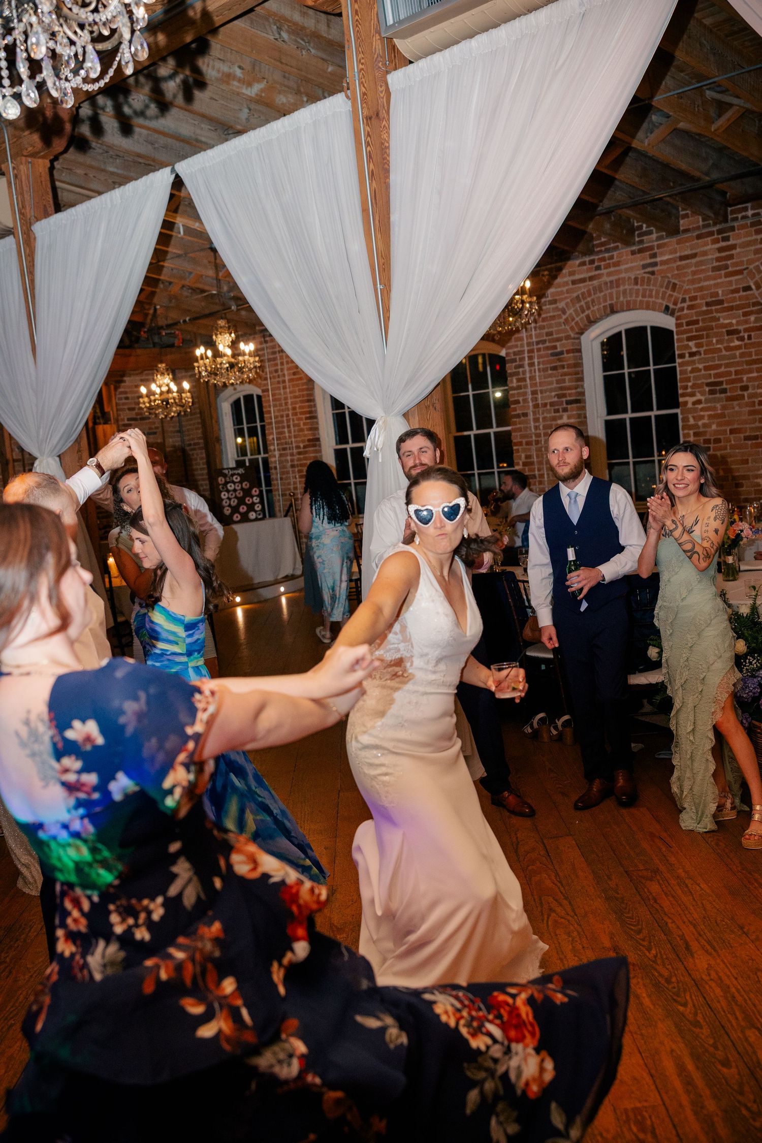Bride wearing fun sunglasses dances energetically with guests at a lively wedding reception.