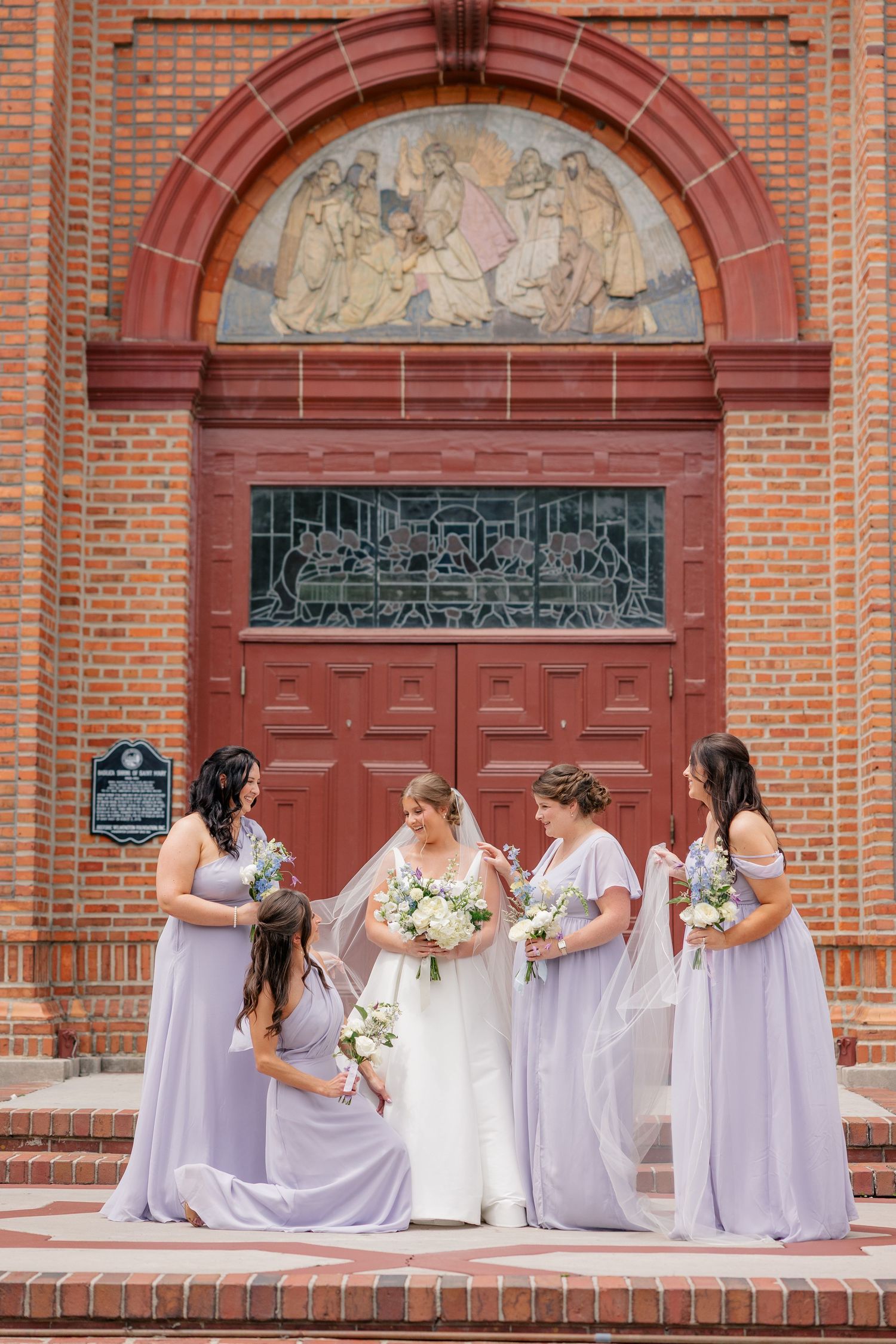 Bride and four bridesmaids in lavender gowns pose on church steps beneath an ornate mosaic archway.