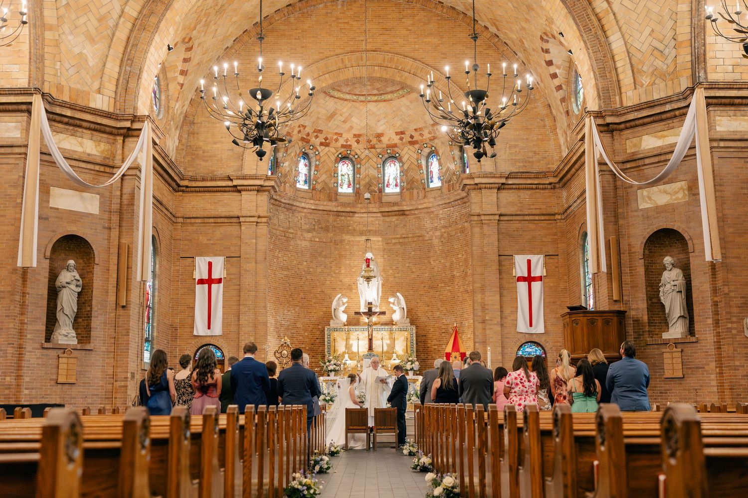Wedding ceremony inside a beautiful brick church with wooden pews, ornate altar, and stained glass windows.