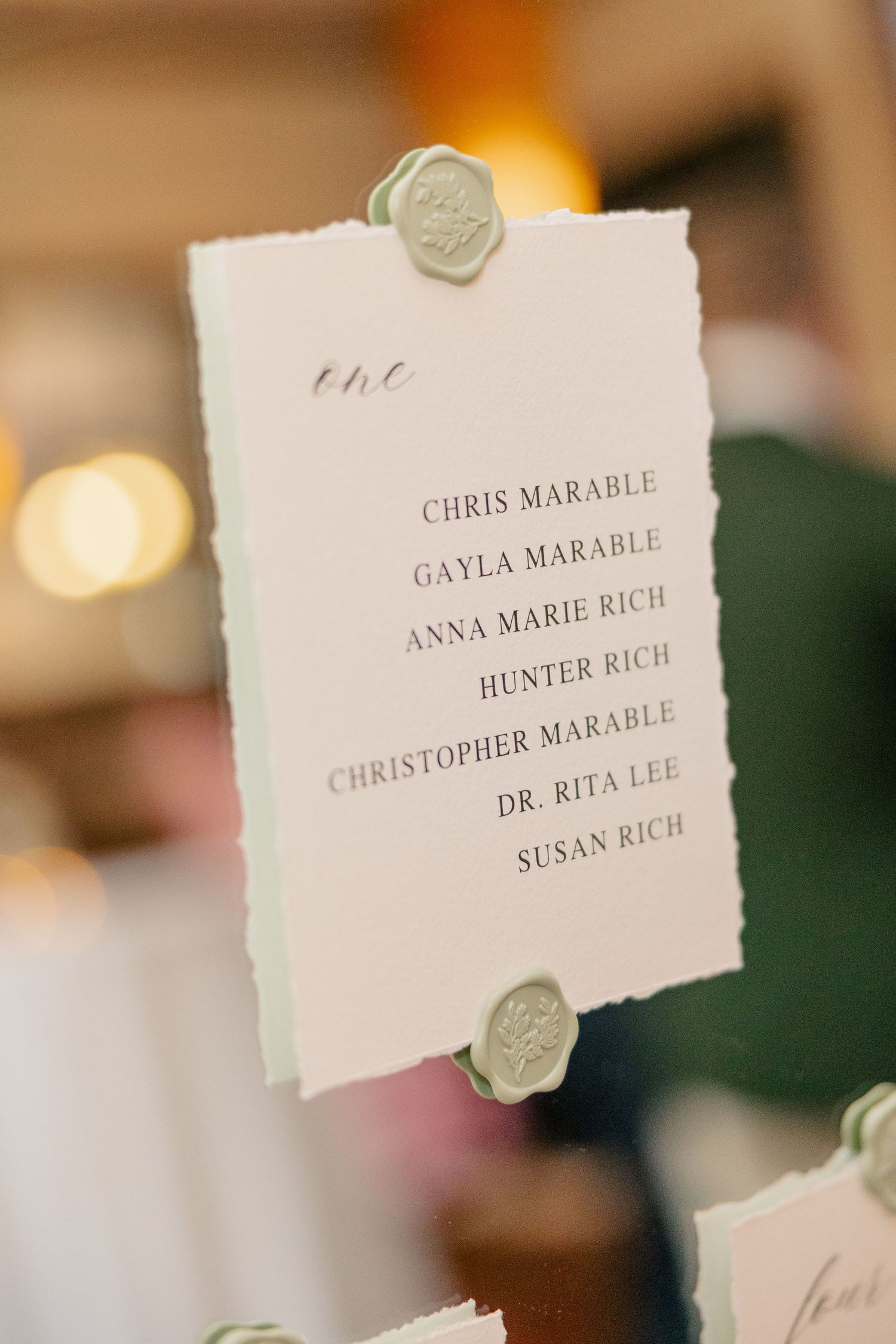 Elegant white wedding seating chart card with green wax seals listing guest names in calligraphy.