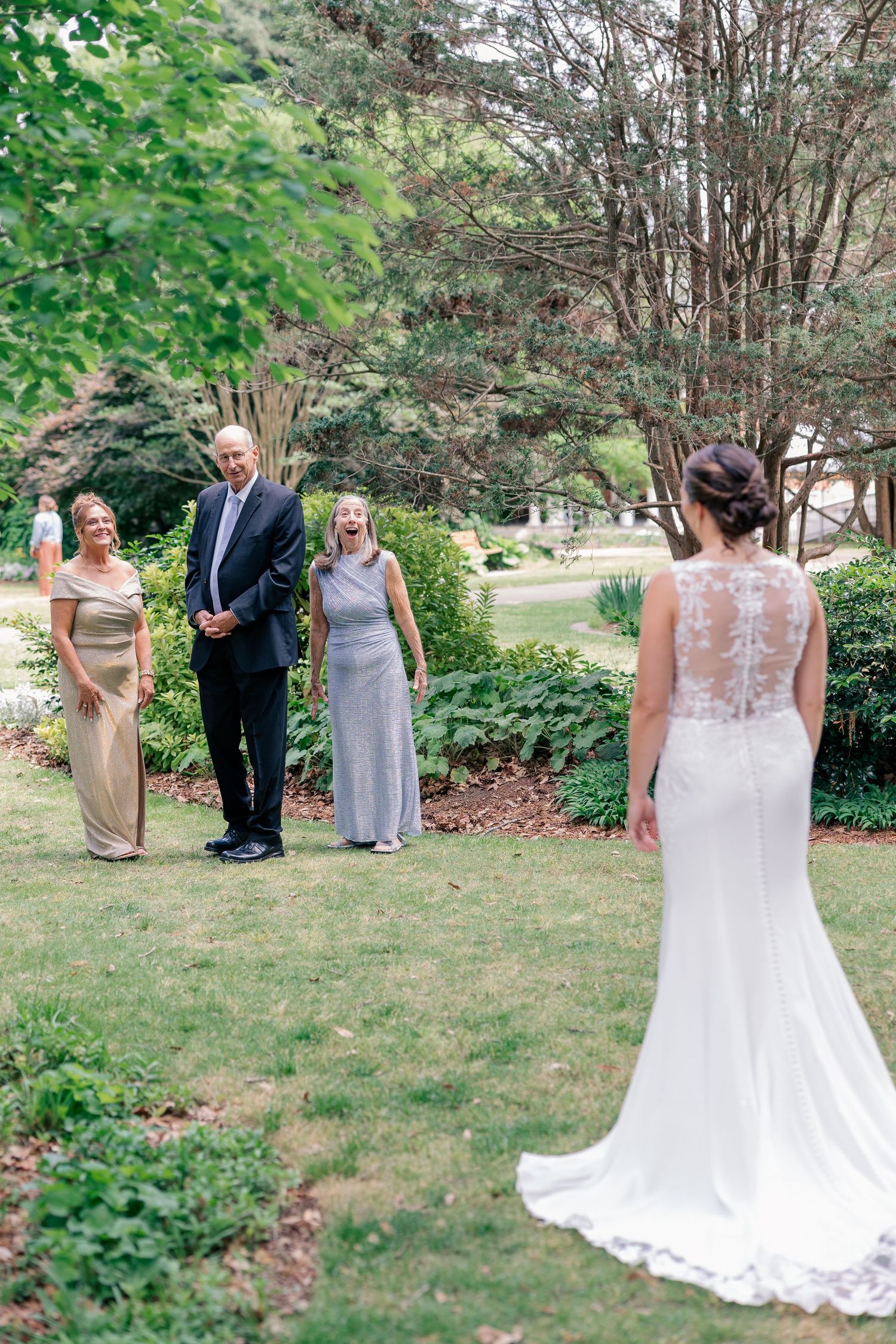 Family reacts with joy seeing bride in lace back wedding dress during outdoor first look moment.