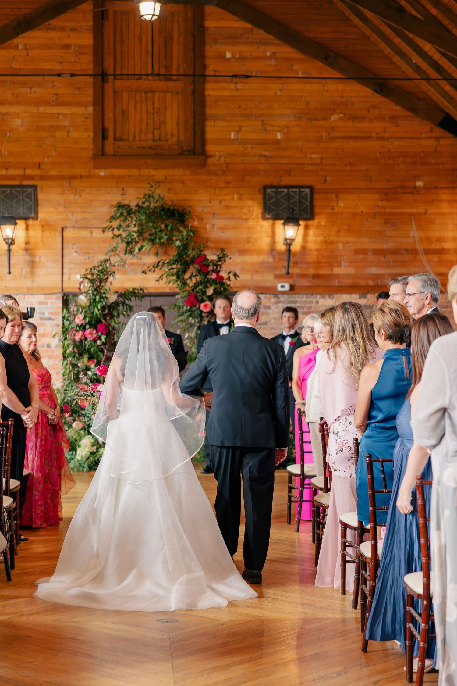 Bride walks down the aisle with escort in rustic wood barn wedding venue with floral arch.