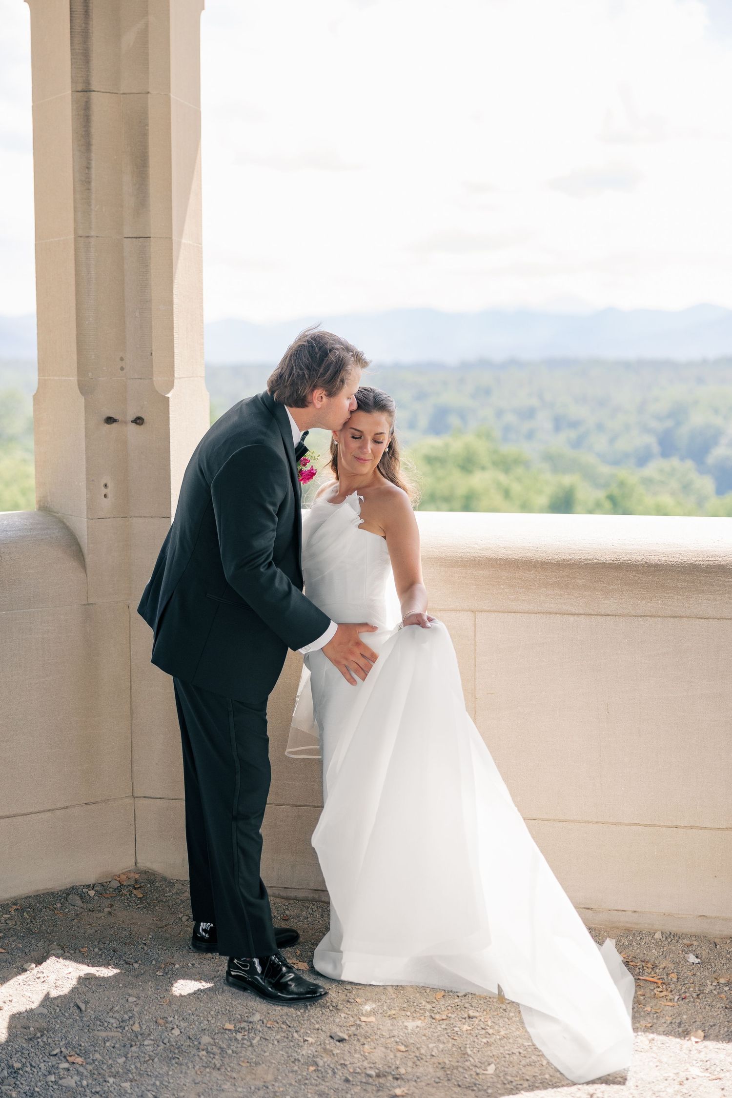 Groom kisses bride on cheek as she smiles on stone balcony with scenic forest backdrop.