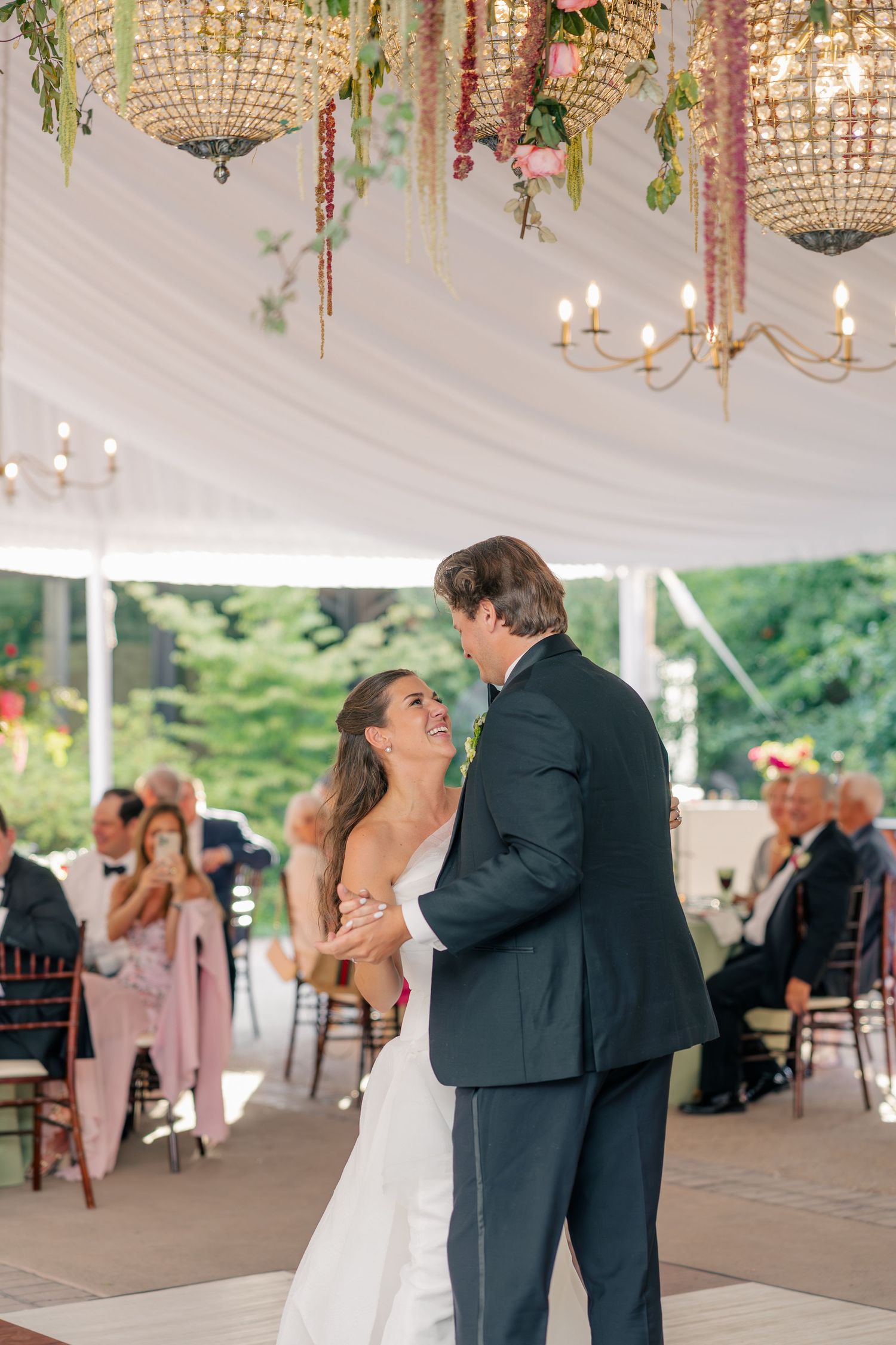 Bride and groom share their first dance under crystal chandeliers adorned with flowers at an elegant tented wedding reception.