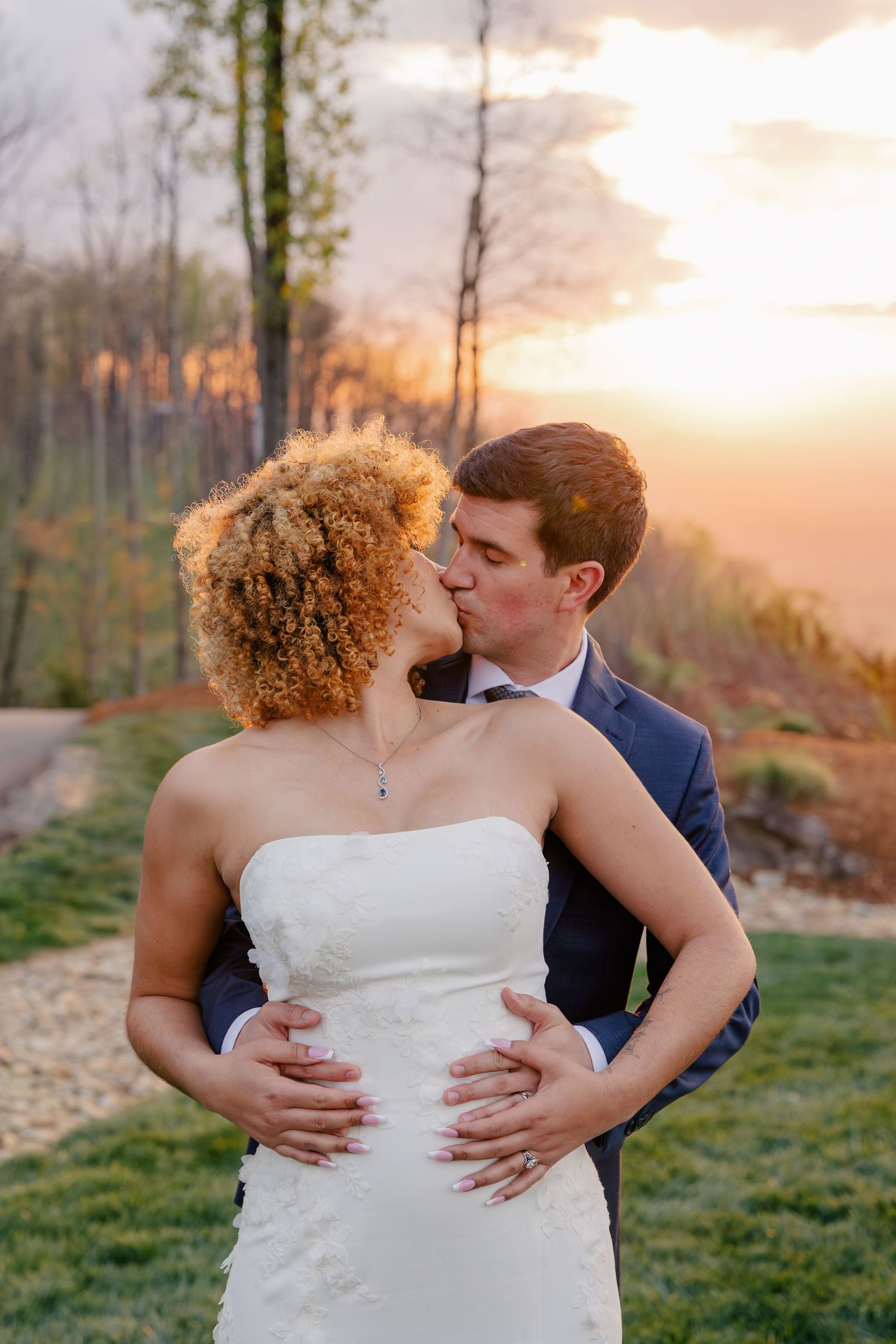 A couple shares a romantic sunset kiss, the bride in a strapless white gown and groom in a navy suit outdoors.