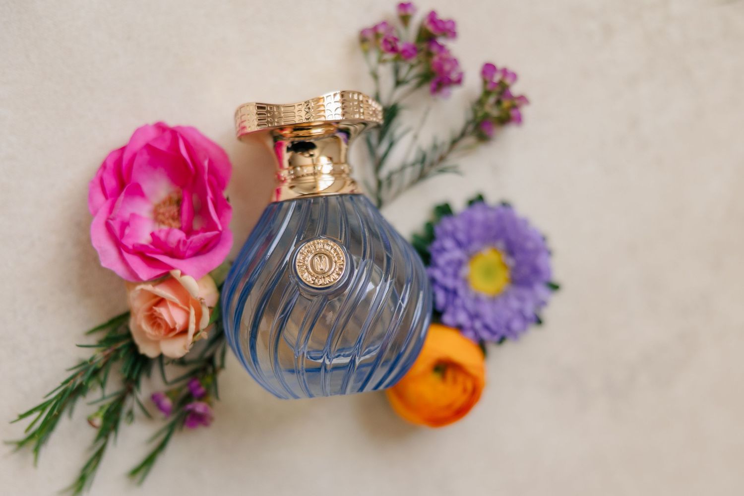 Blue and gold perfume bottle surrounded by colorful pink, purple, and orange flowers on white background.