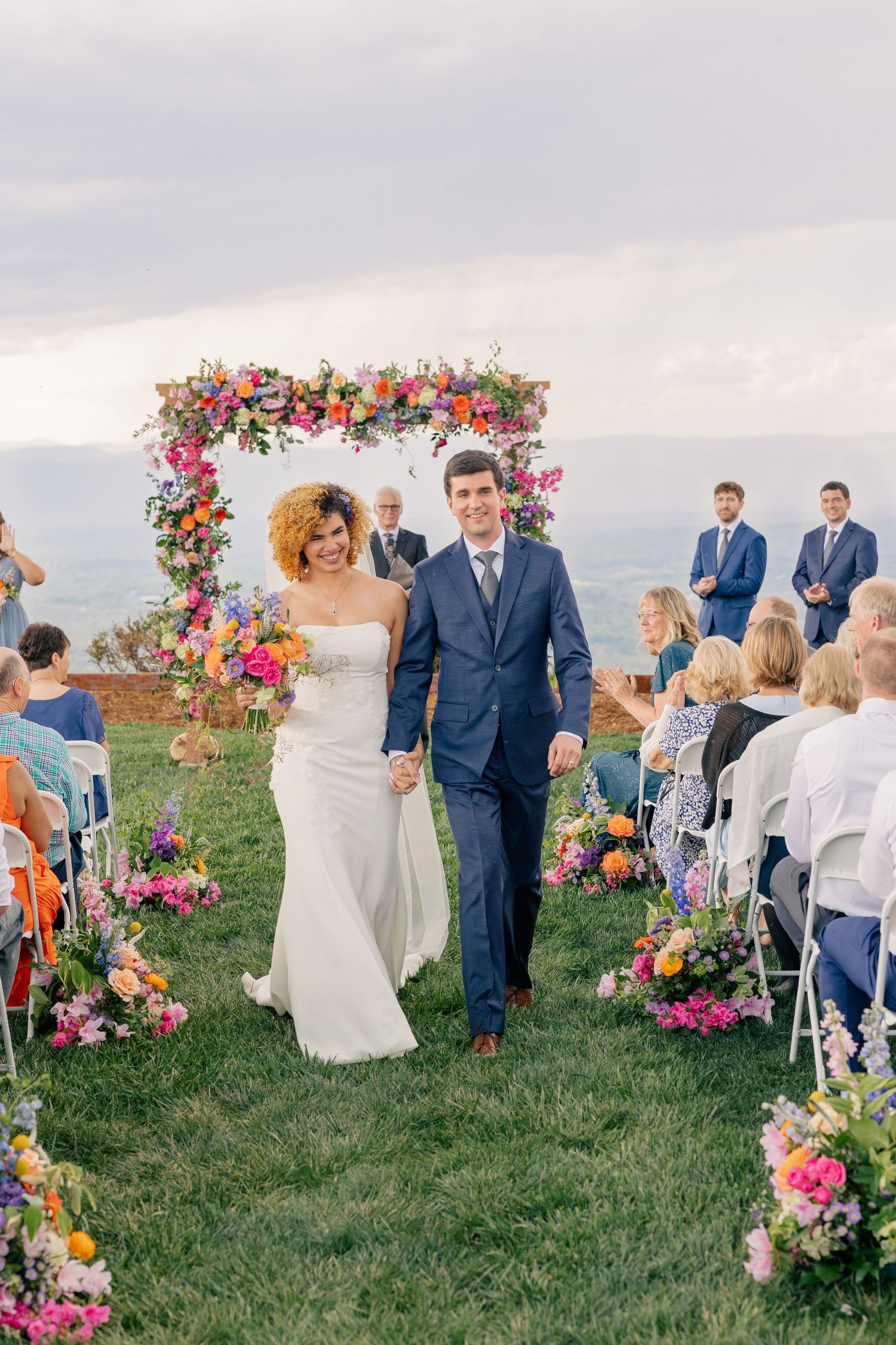 Newlyweds walking down aisle after ceremony under colorful floral arch with guests applauding outdoors.