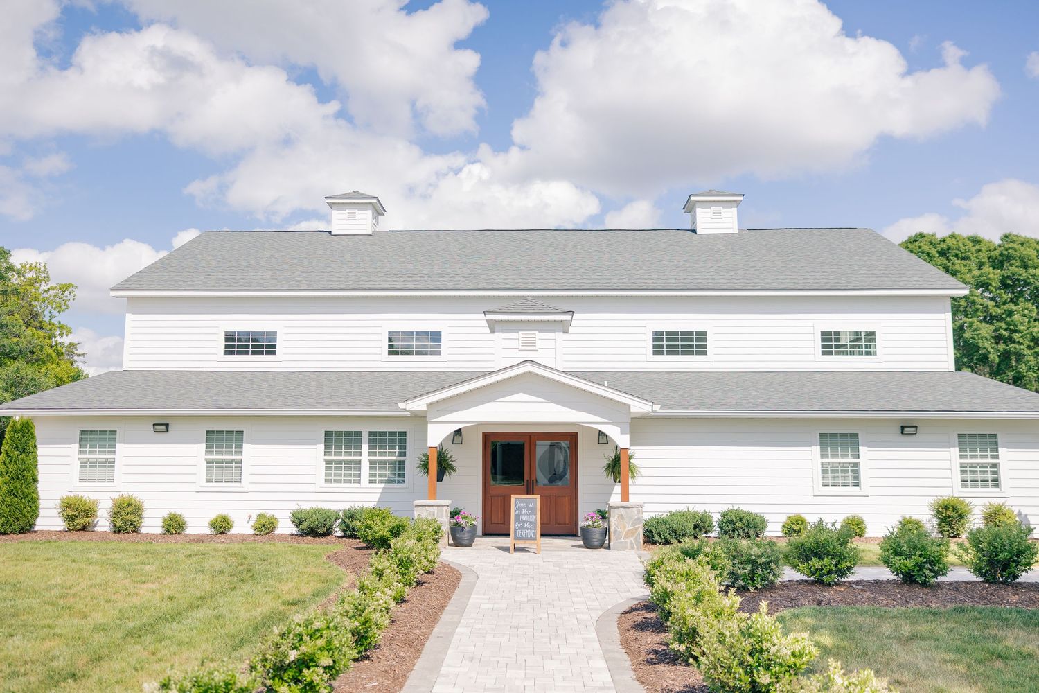 Large white farmhouse with gray roof, manicured shrubs, stone pathway, and wooden front door on a sunny day.