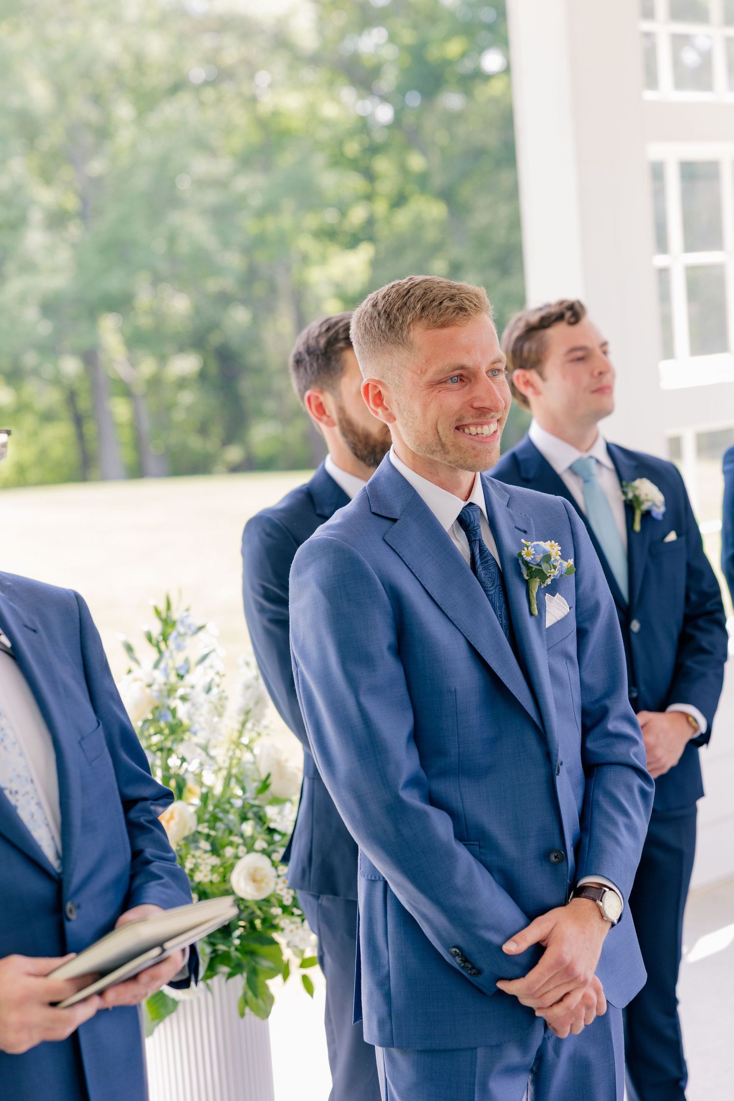 A smiling groom in a blue suit watches his bride walk down the aisle during an outdoor wedding ceremony.