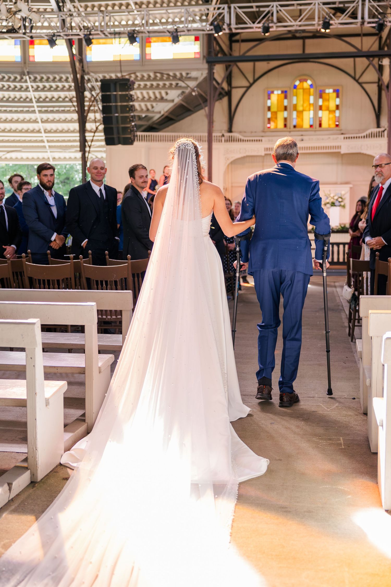 Bride in flowing white gown and veil walks down the aisle with escort in blue suit at church ceremony.