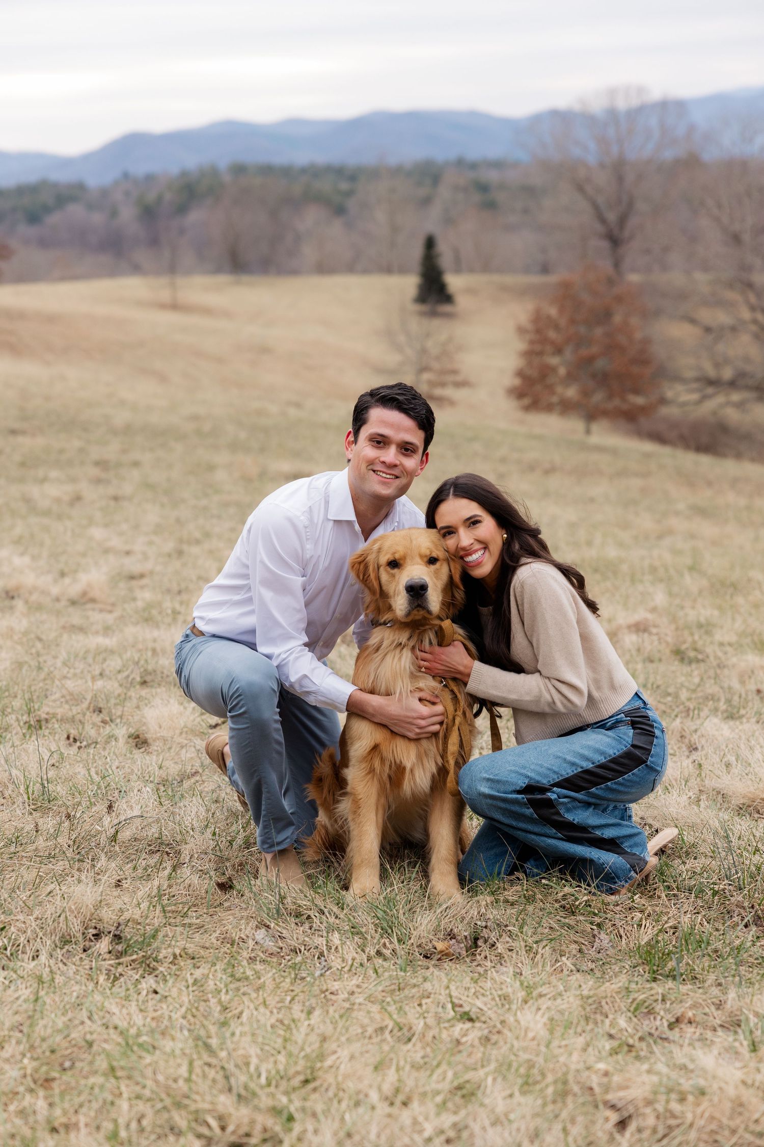 A couple kneels with their Golden Retriever in a grassy field with mountains in the background during fall.