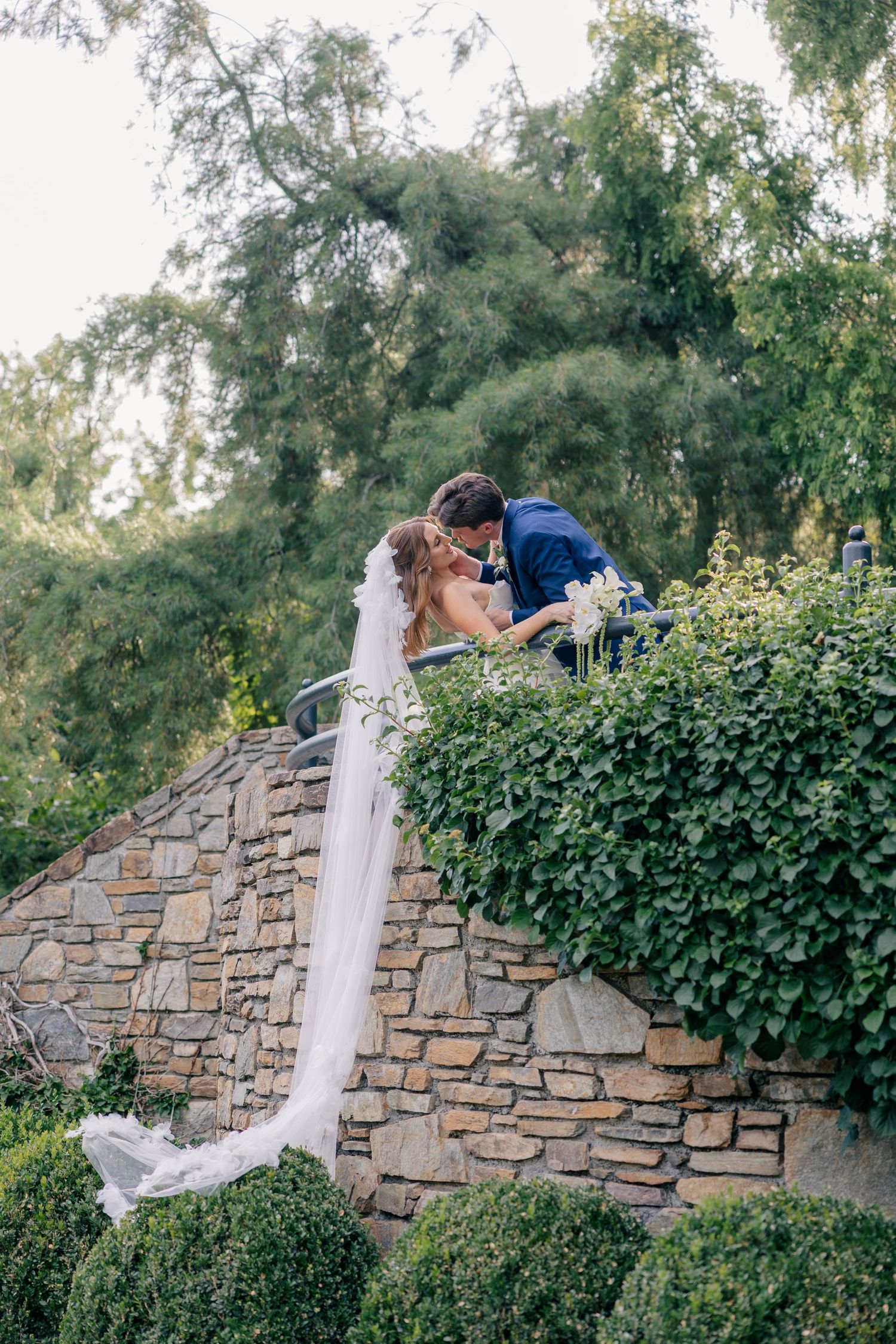 A romantic wedding couple shares intimate moments against a rustic stone wall backdrop in a garden setting. Arboretum Wedding Photographer