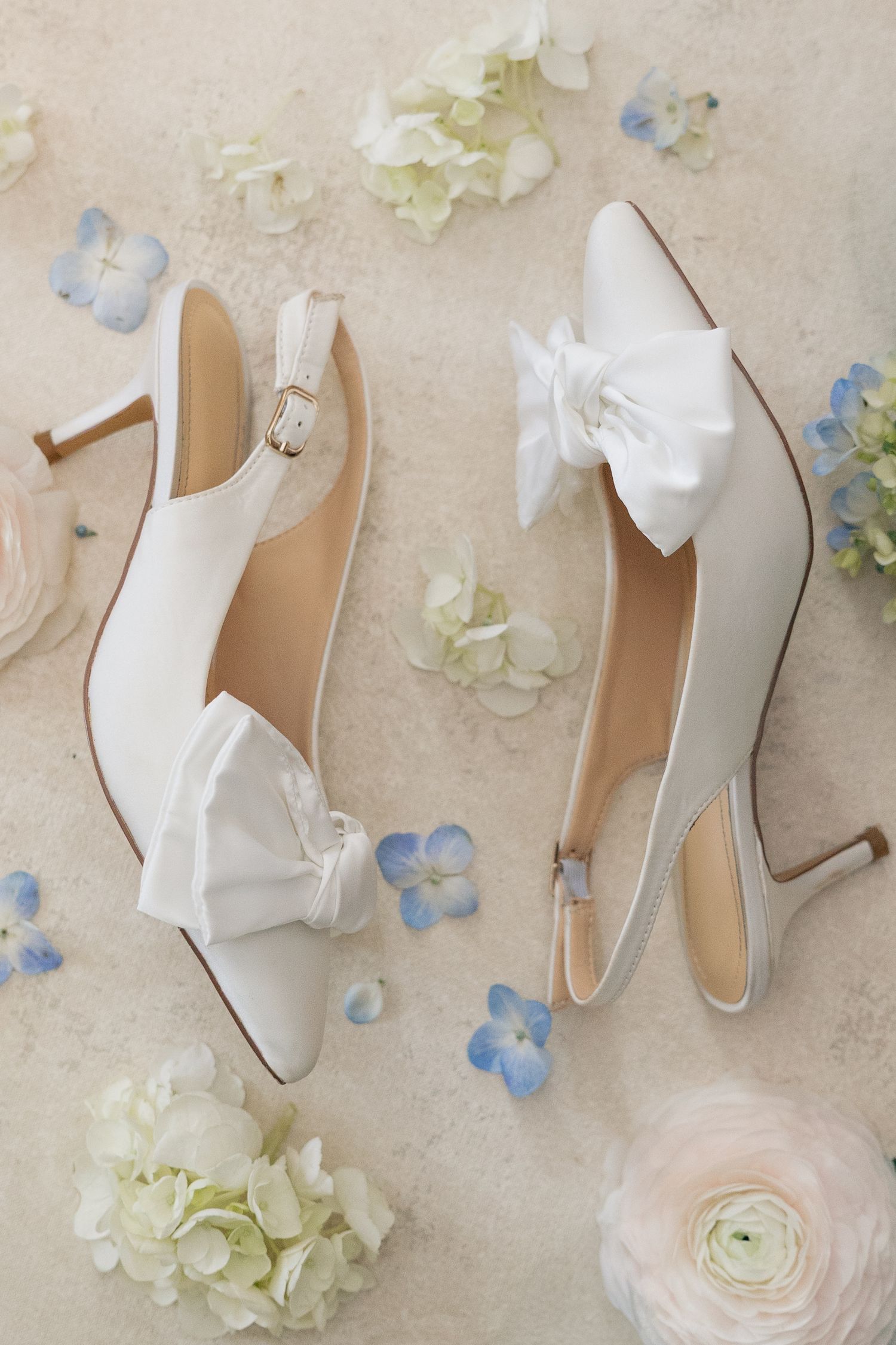 White satin bridal heels with bow detail surrounded by blue hydrangeas and pink ranunculus flowers on a neutral background.