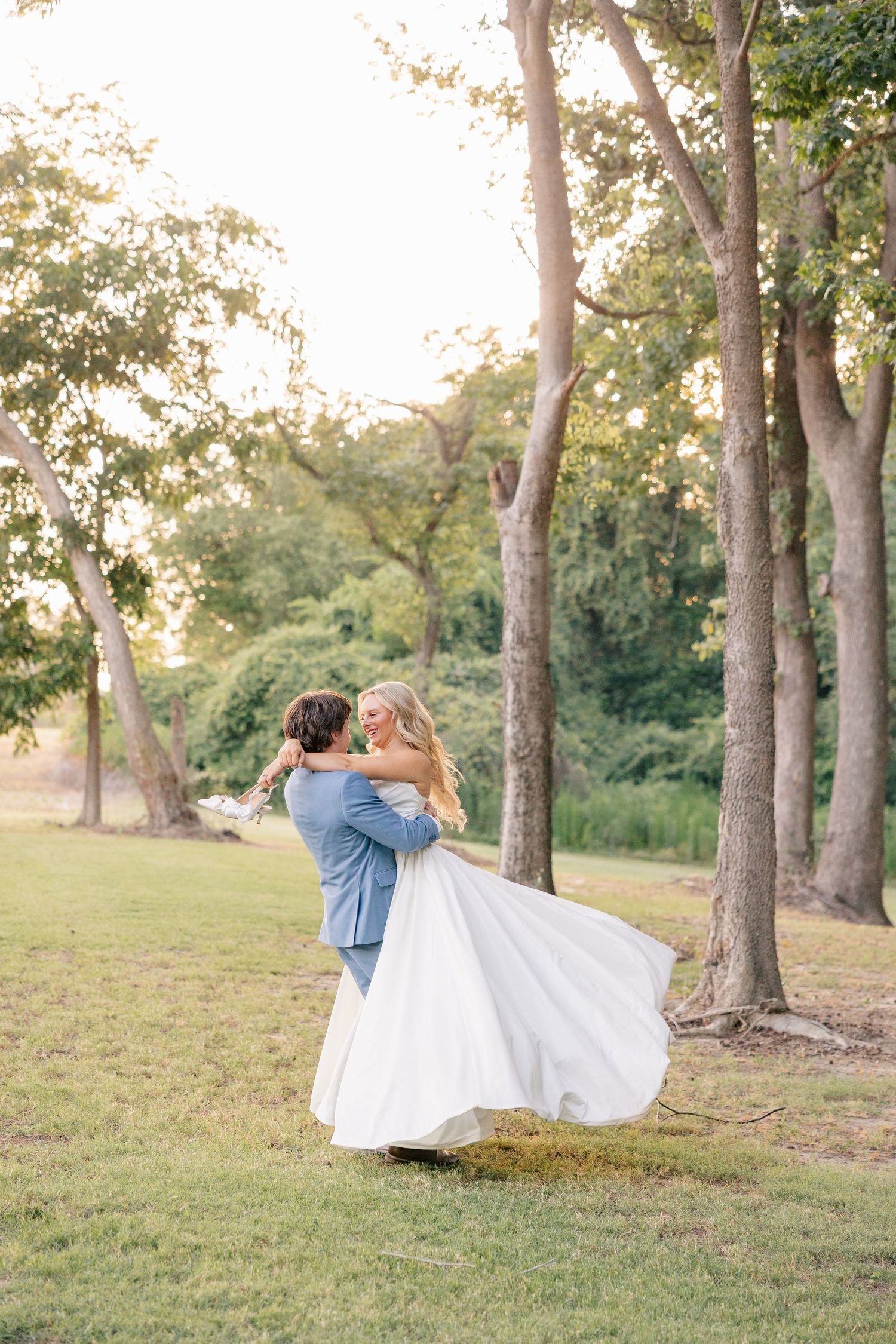 Groom in blue suit lifts and spins bride in white gown among golden sunlit trees.