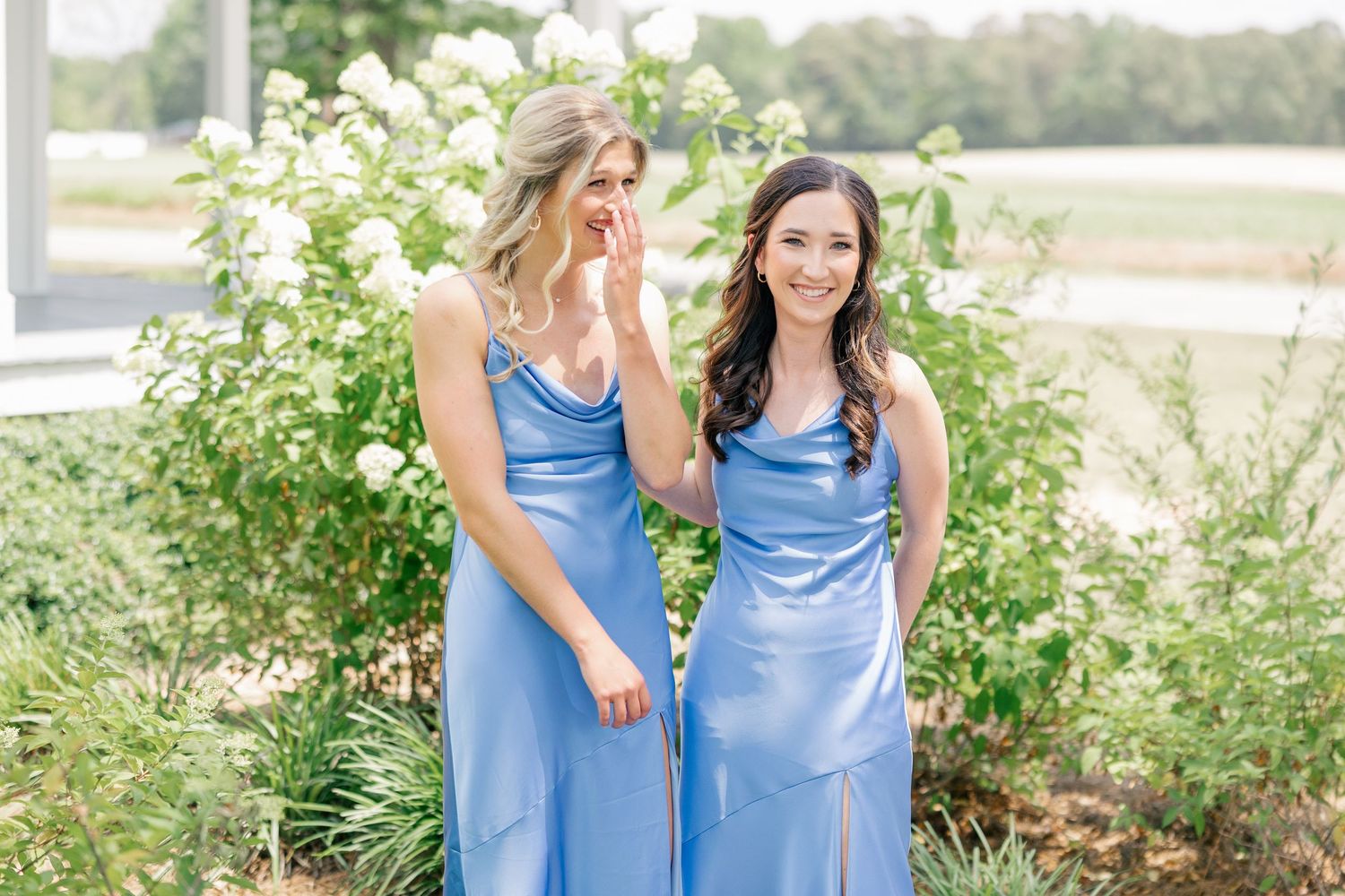 Two bridesmaids in periwinkle blue satin dresses laughing in front of white roses.