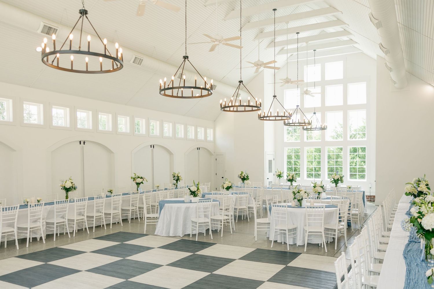 Elegant white wedding reception hall with checkered floors, round chandeliers, and white chiavari chairs.