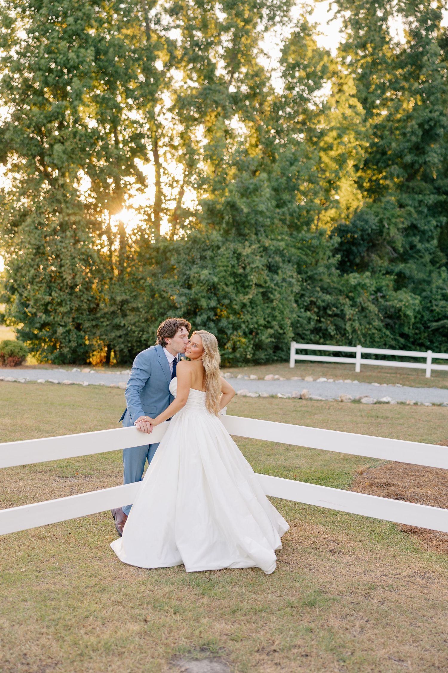 Bride and groom sharing a tender kiss near a white fence at sunset on their wedding day.