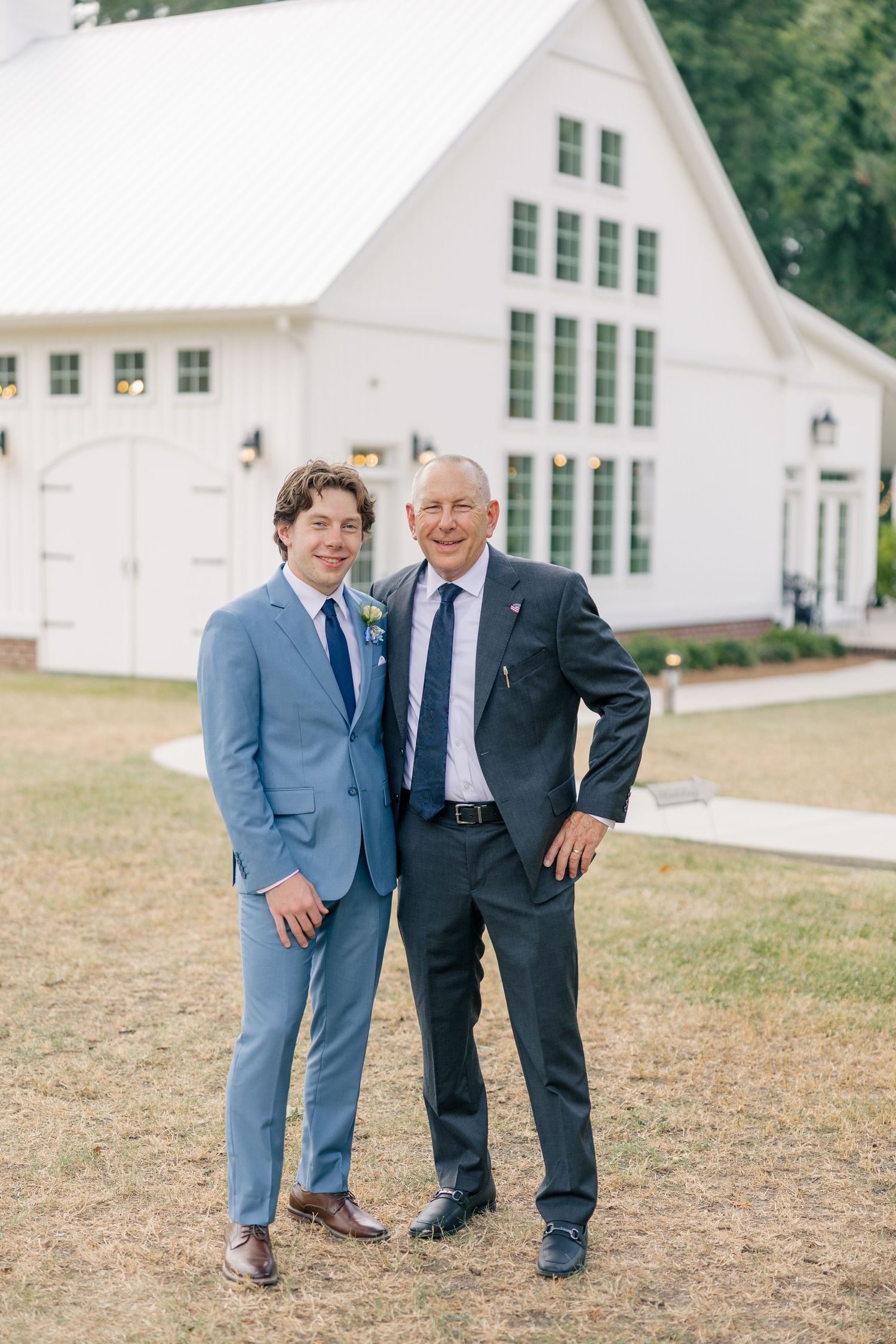 Groom in light blue suit poses with older gentleman in navy suit outside white barn.