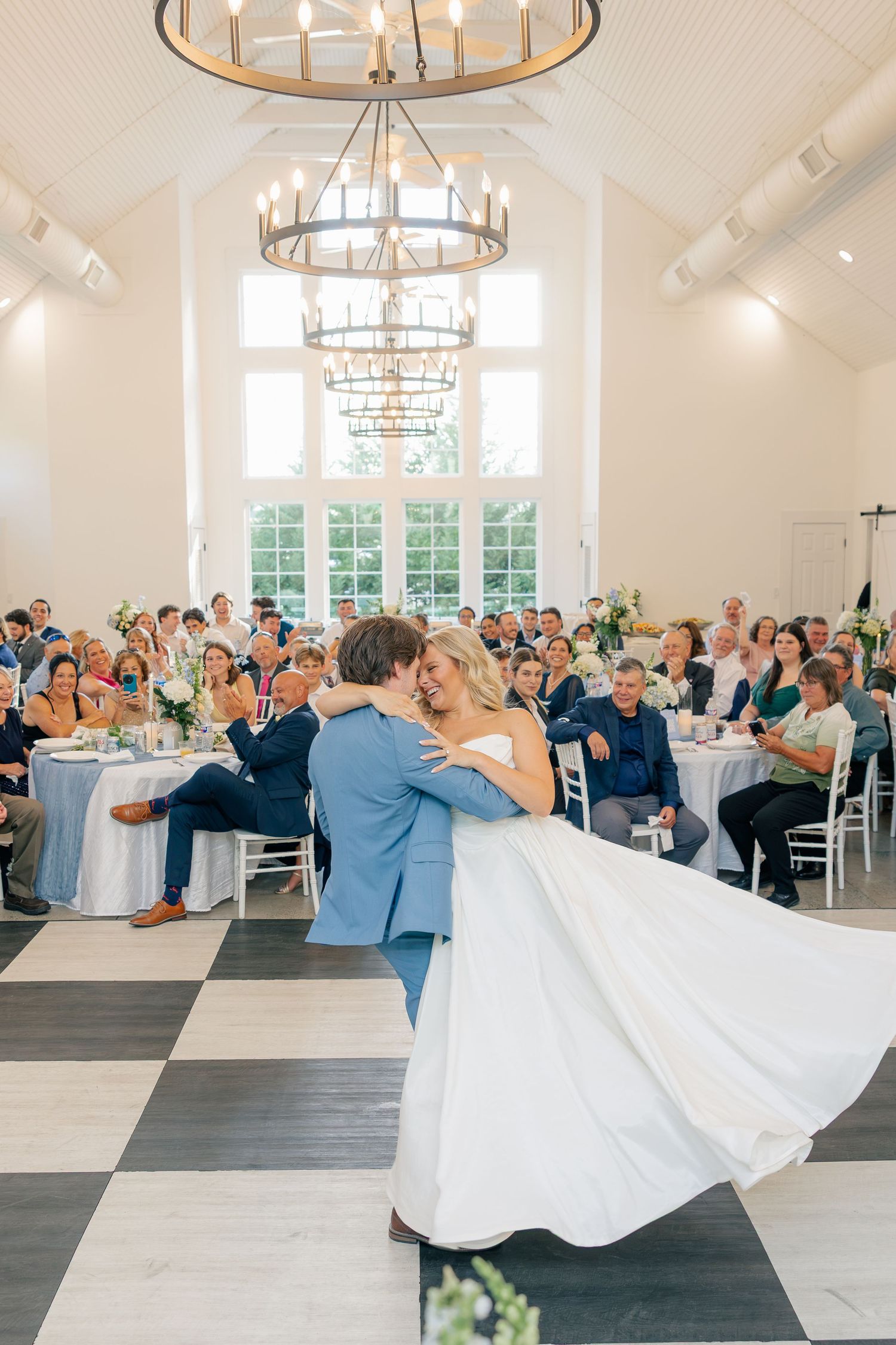 Bride and groom share first dance in elegant white reception hall with chandelier and checkered dance floor.