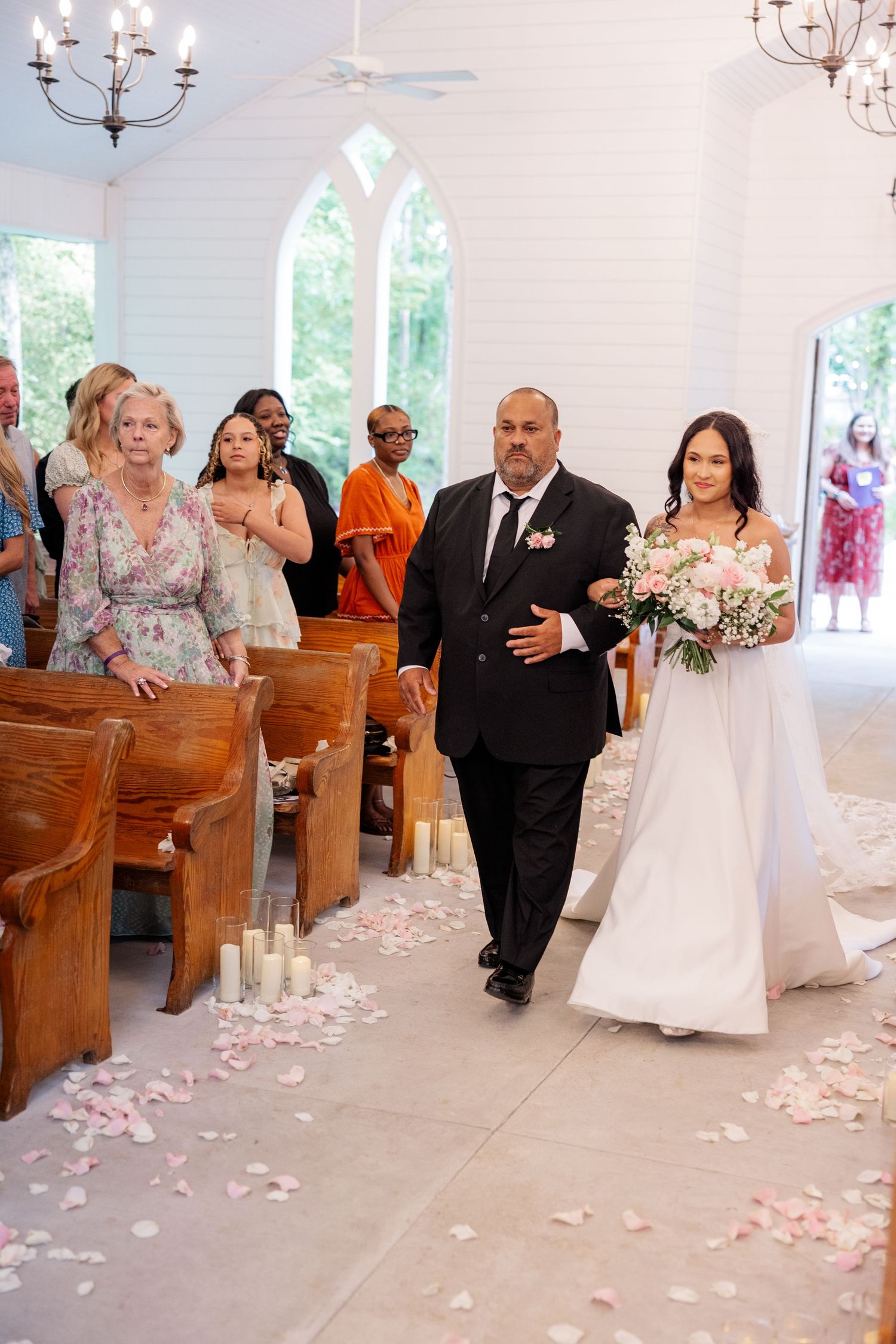 Wedding party walks down church aisle with pink rose petals scattered on floor as guests look on from wooden pews.
