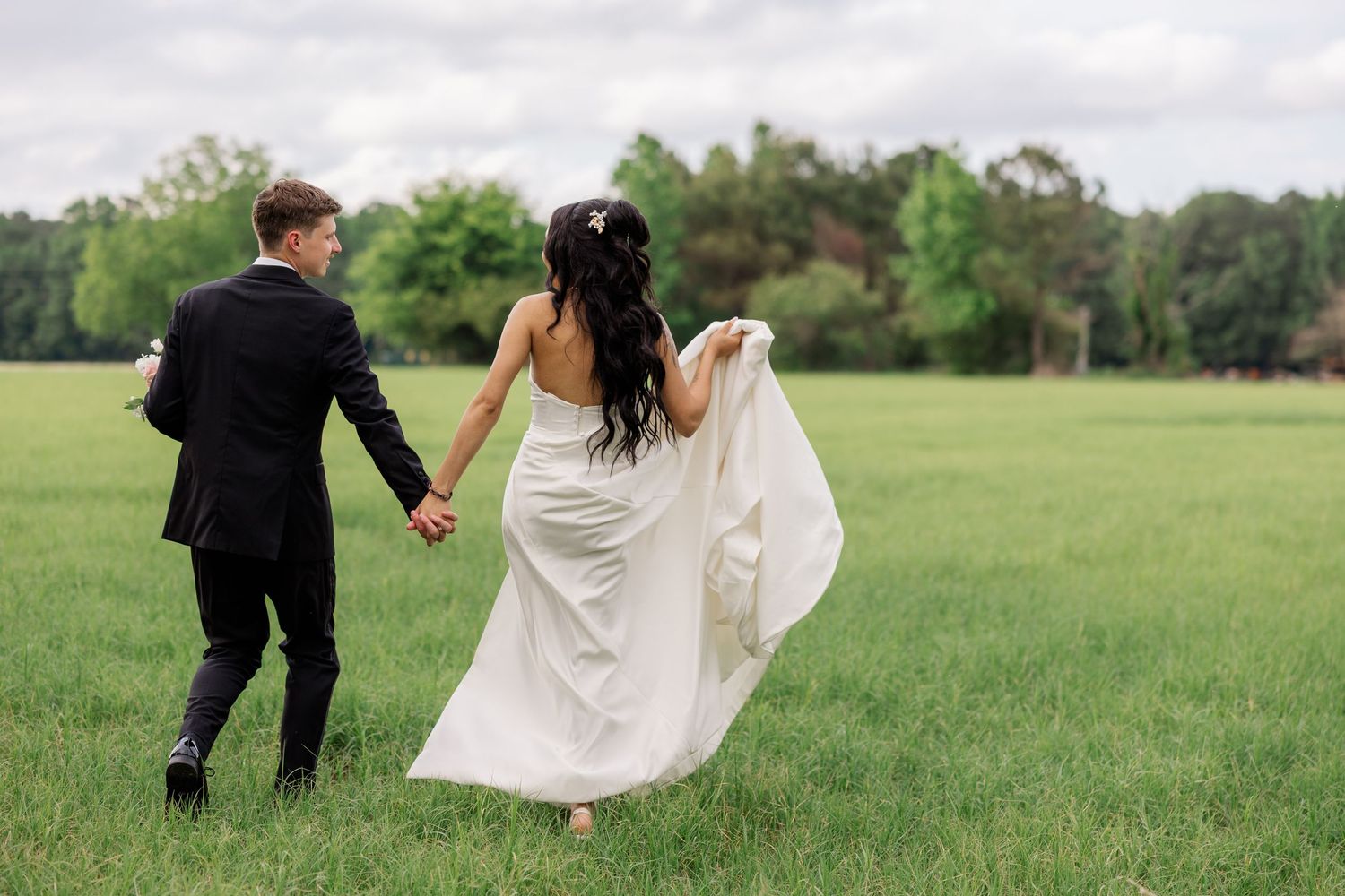 Romantic wedding couple walking through green field with flowing white dress and black tuxedo.