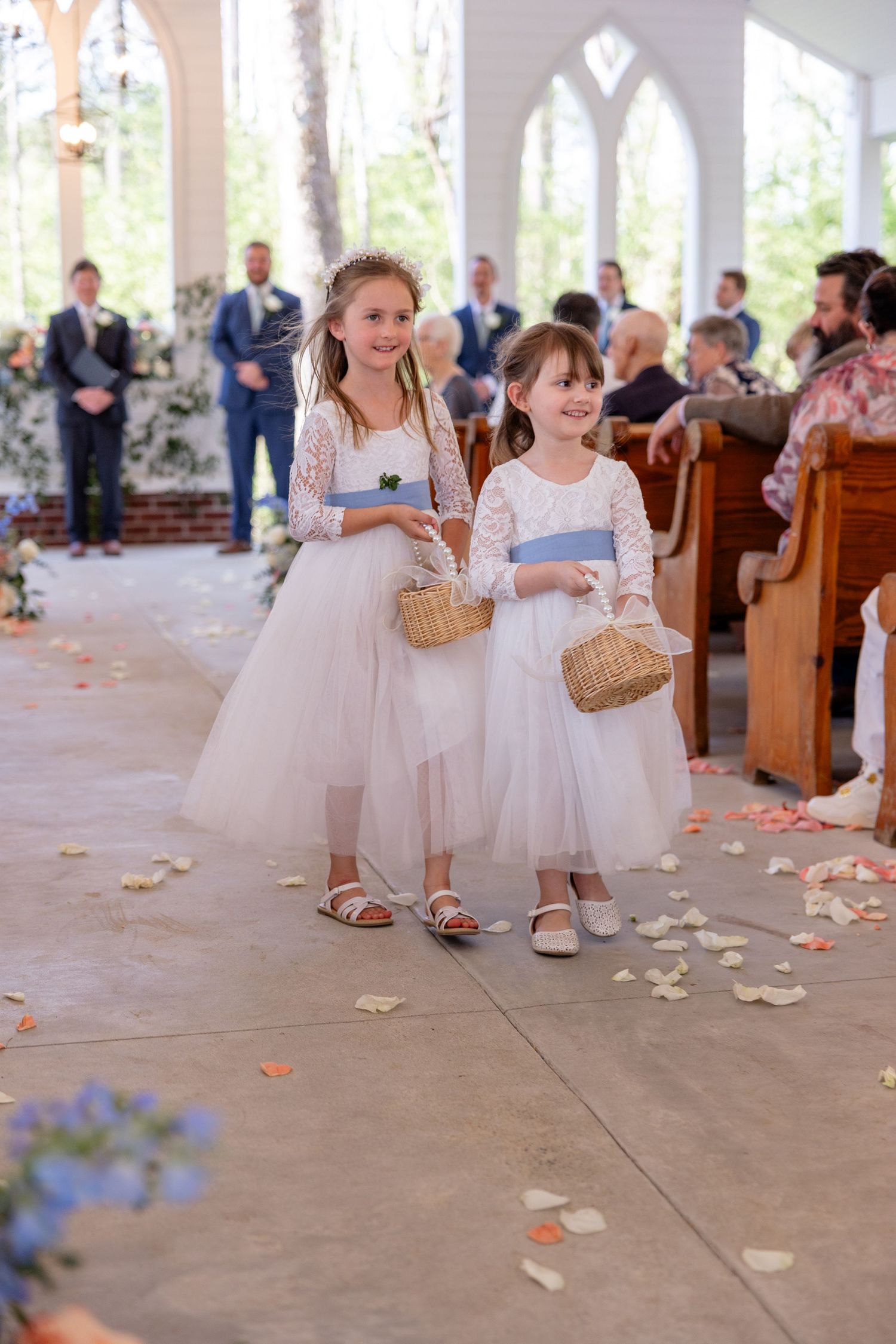 Young flower girls in white dresses scatter rose petals down church aisle during wedding ceremony.