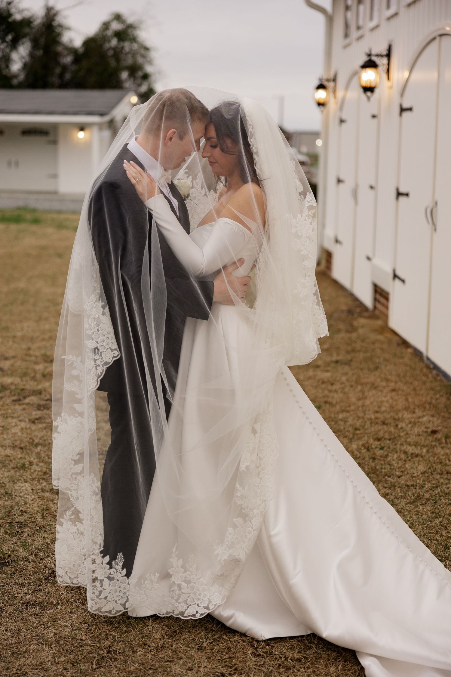 Romantic outdoor wedding kiss captured against white barn backdrop with string lights.