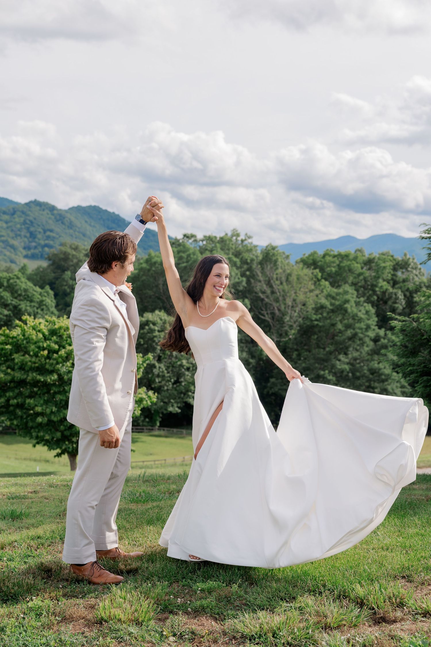 A couple dancing outdoors in wedding attire with a flowing white dress against a mountain backdrop.