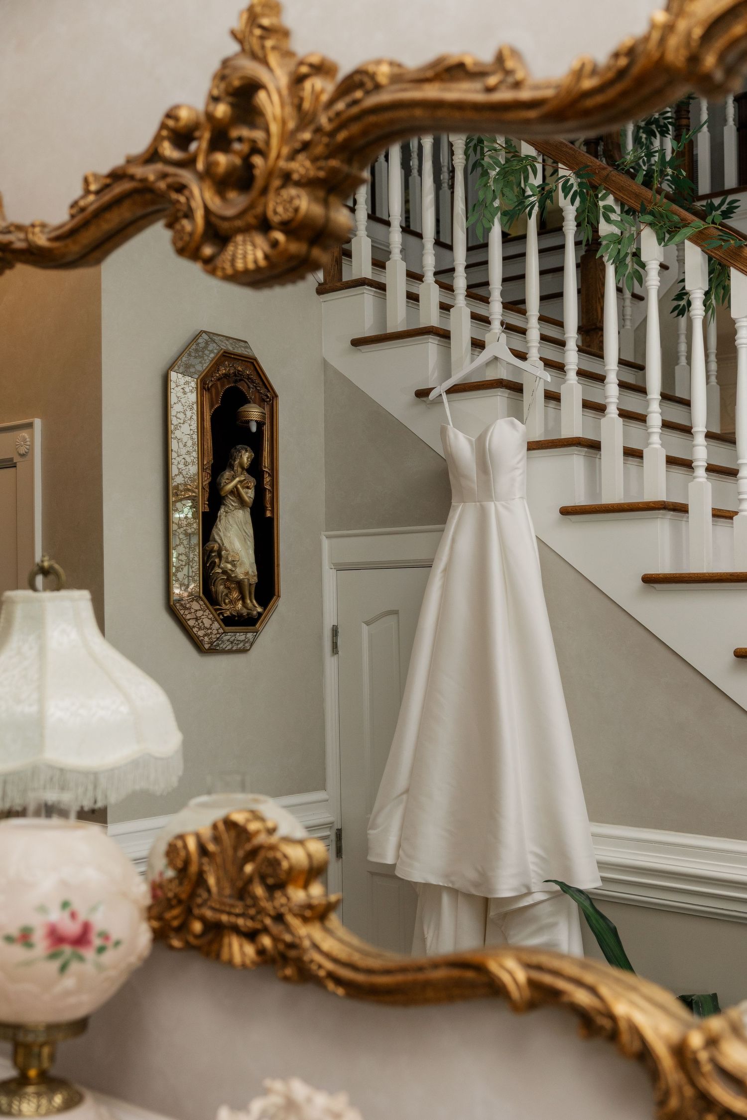 Elegant white wedding dress hangs from ornate gold-framed mirror against white staircase in luxurious interior setting.