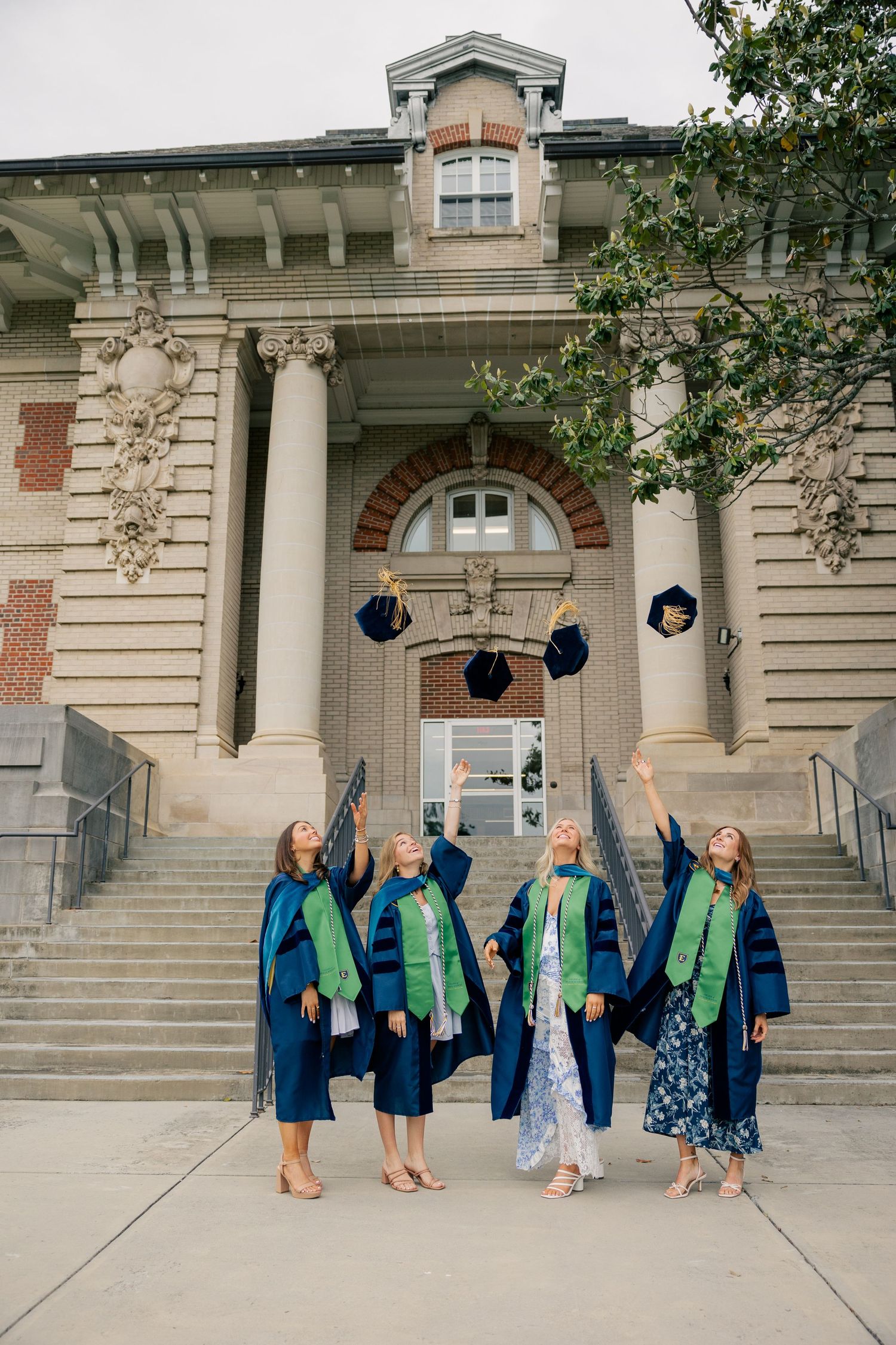 Graduates in blue caps and gowns throw their caps in celebration on stone steps of historic university building.