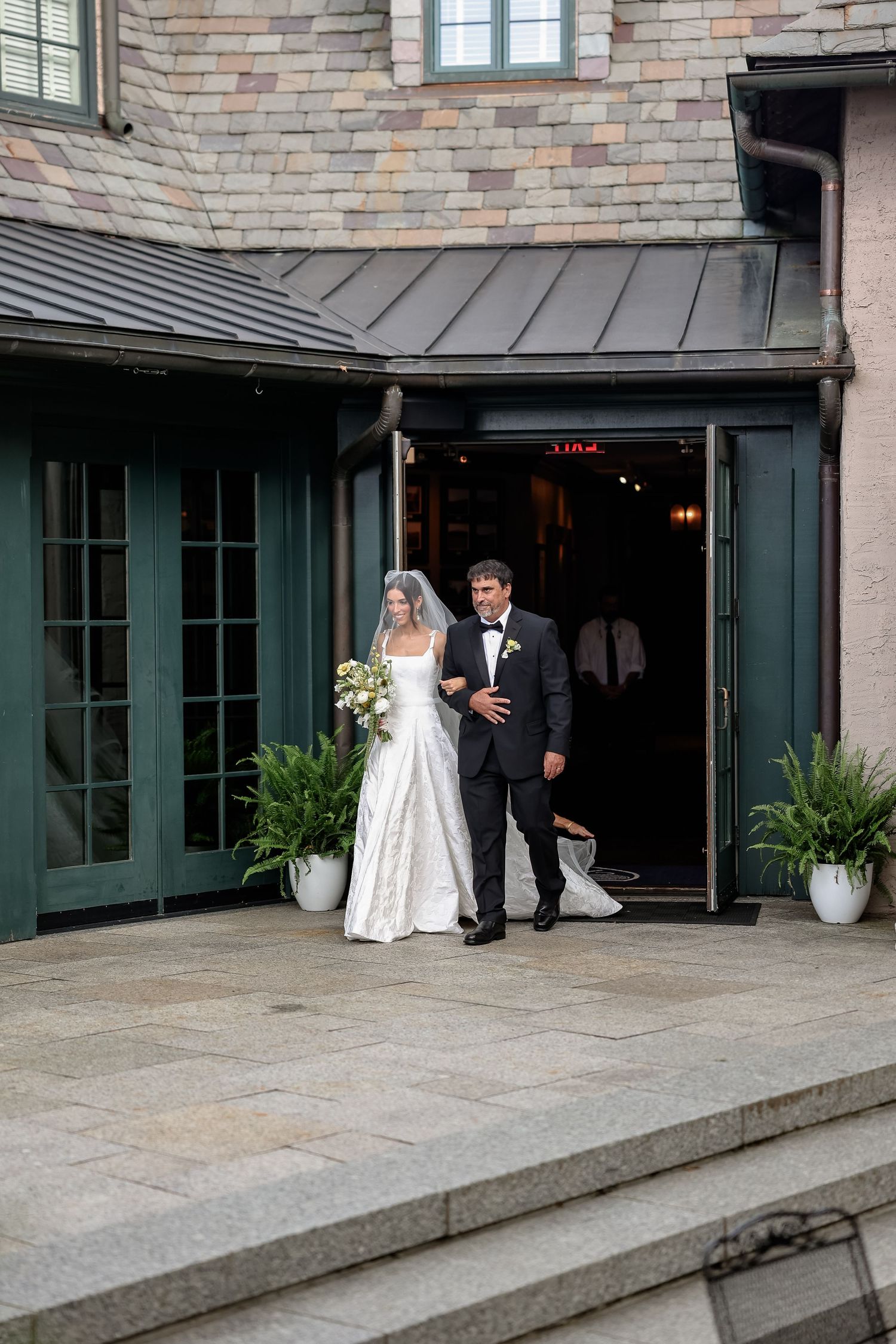 Bride in white gown and veil walks with escort in tuxedo out of elegant stone venue entrance.