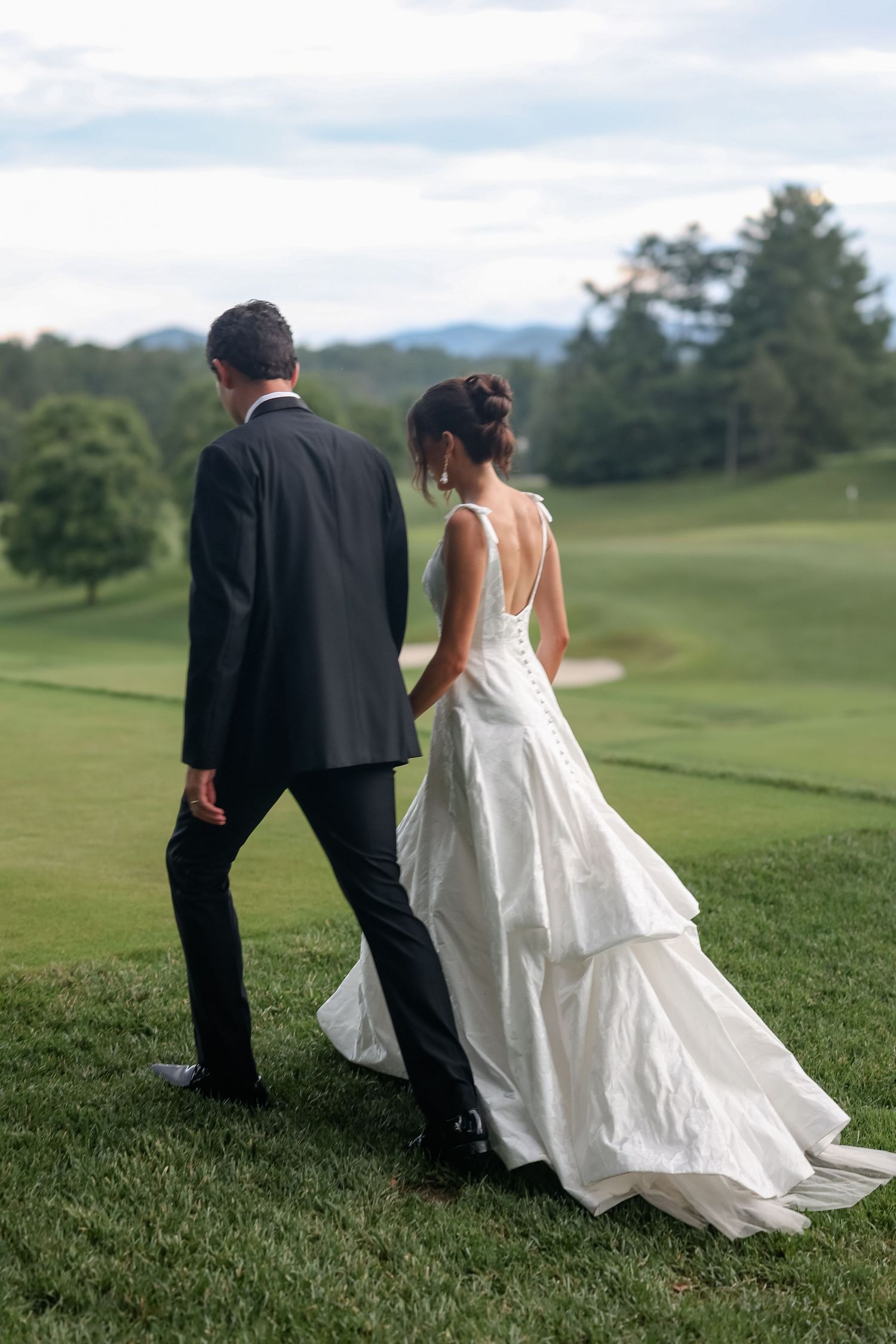 Bride and groom walk hand-in-hand across lush green golf course at dusk after wedding ceremony.